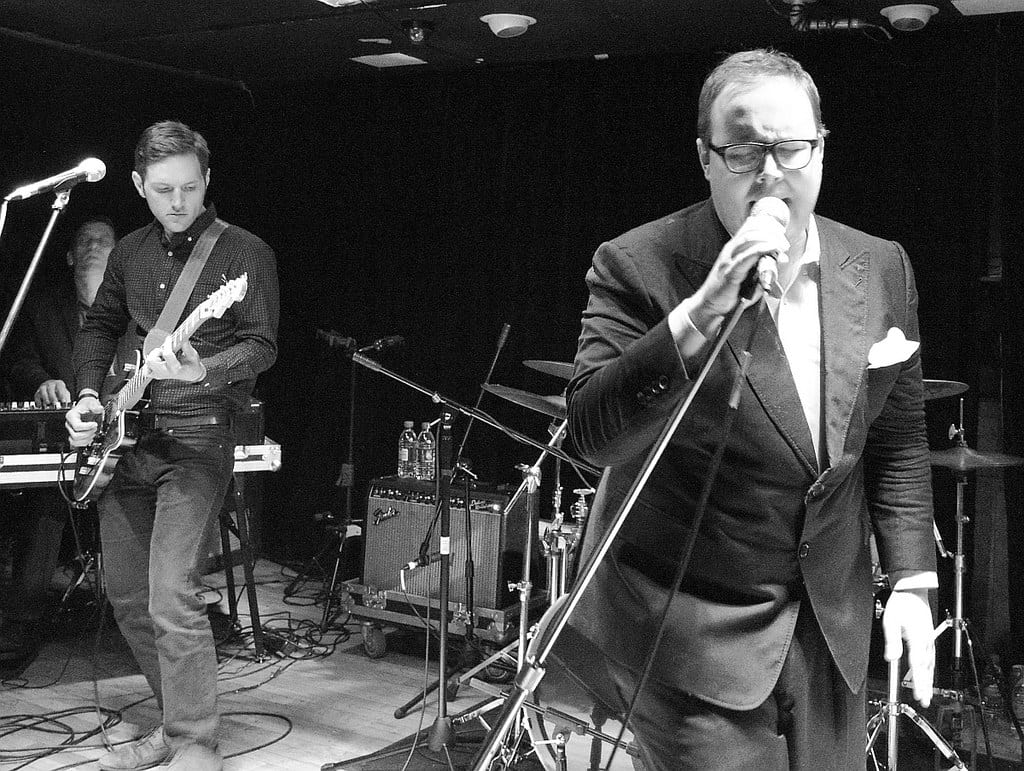 Black and white of a band with a singer in the foreground along with the guitarist and keyboard player in the background