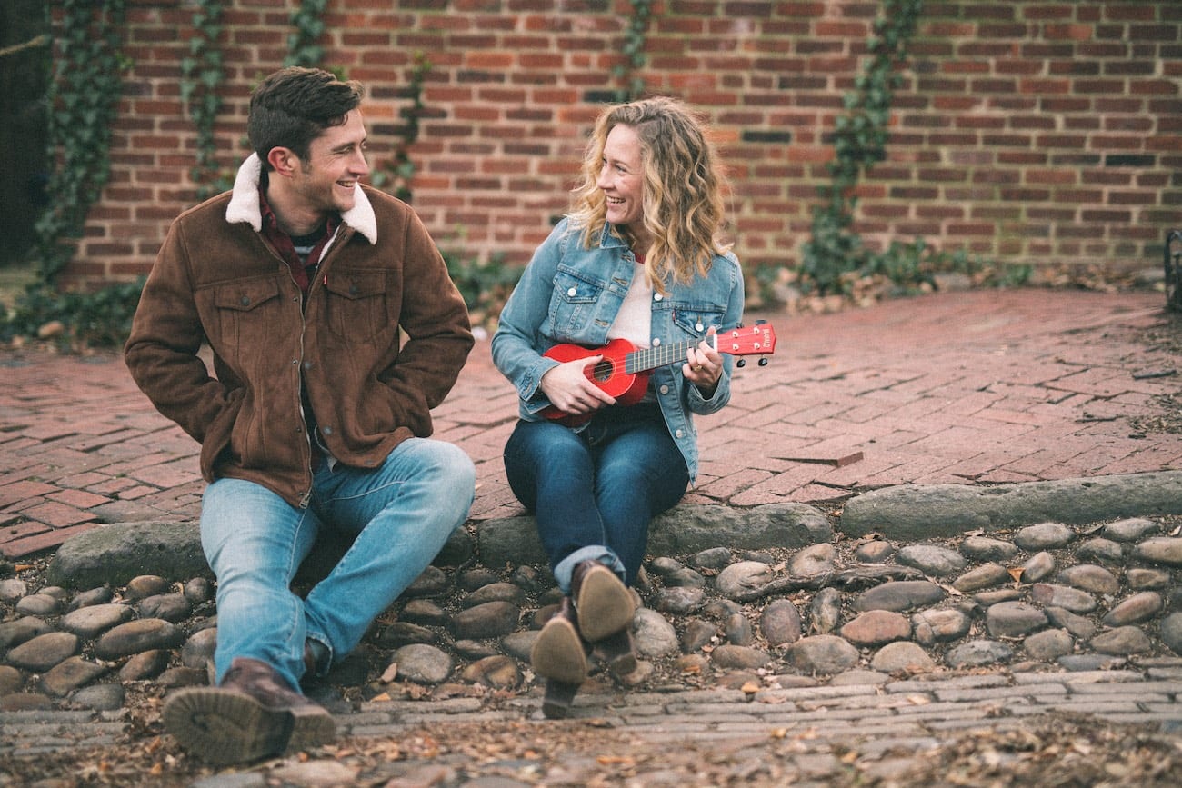 Man and woman with small red guitar sitting on a curb talking to each other