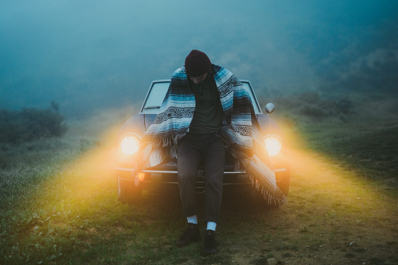 Salma El Wardany Uncreative Poetry C&I Studios Male posing looking down in front of an old car with headlights on wearing an American Indian themed blanket over his shoulders.