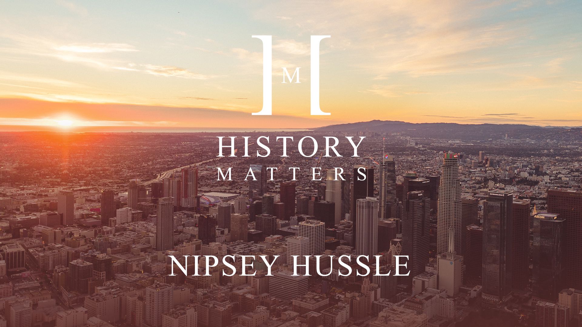 White HM Nipsey Hussle logo with a background of aerial view of a city at sunrise