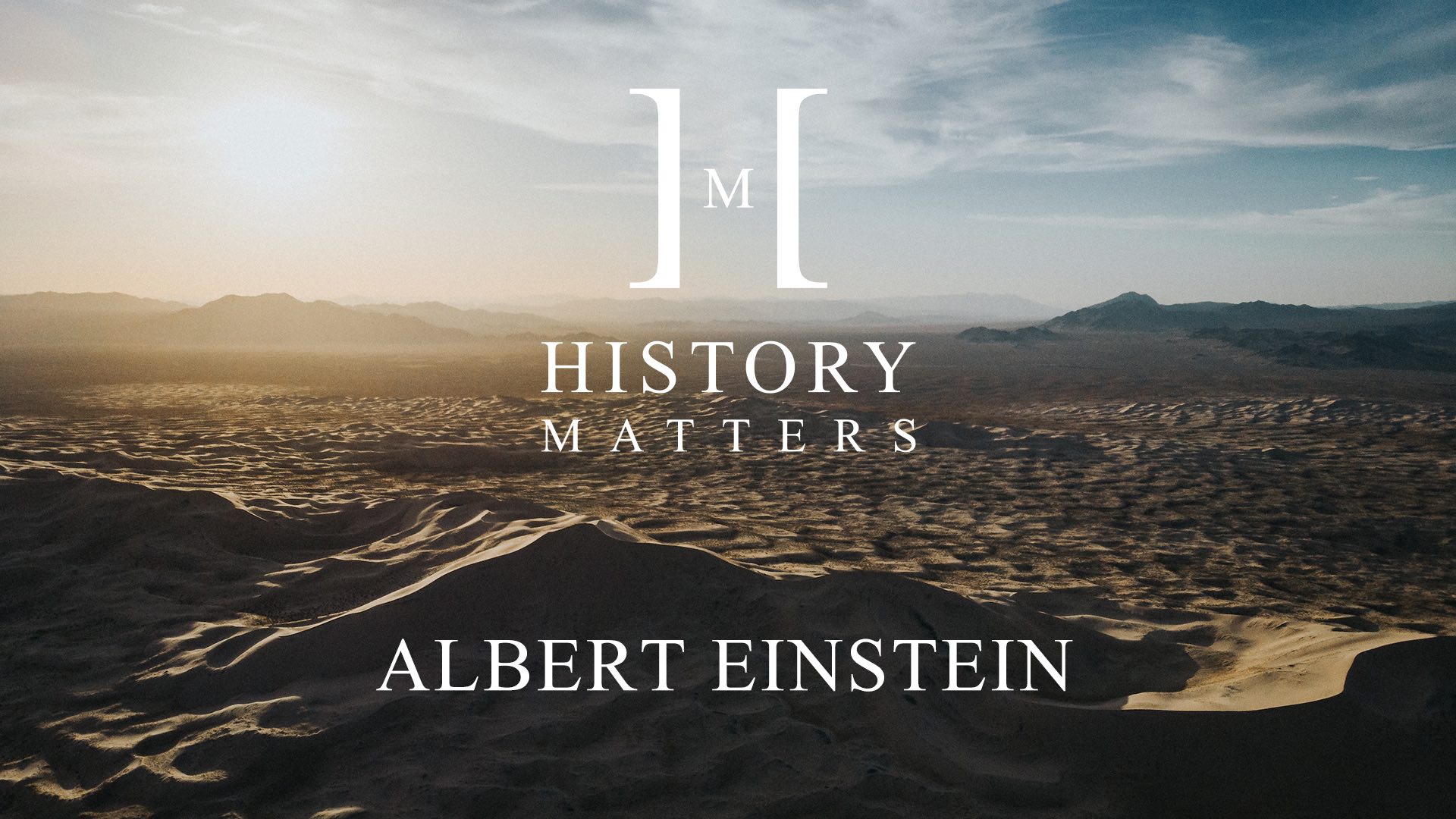 White HM Albert Einstein logo with background of sand dunes and mountains in a desert