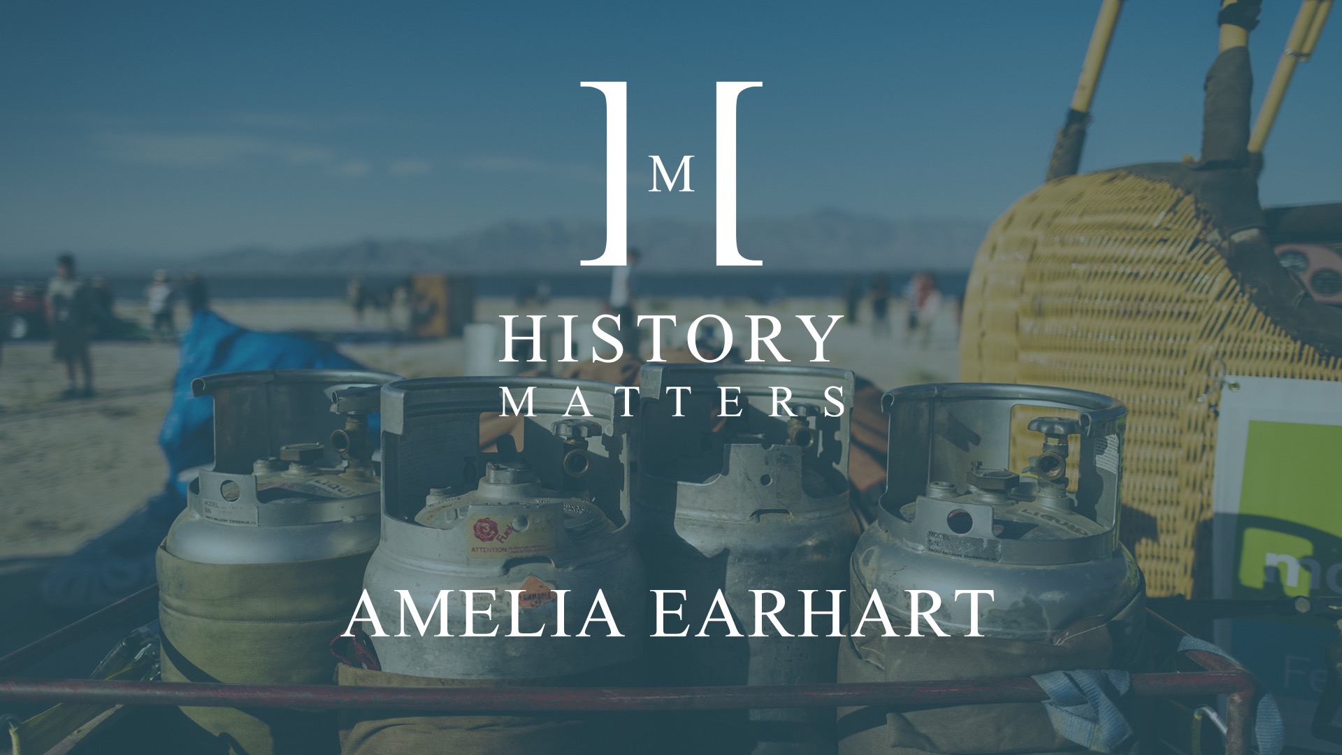 White HM Amelia Earhart logo with background of four fuel metal cannisters by a hot air balloon