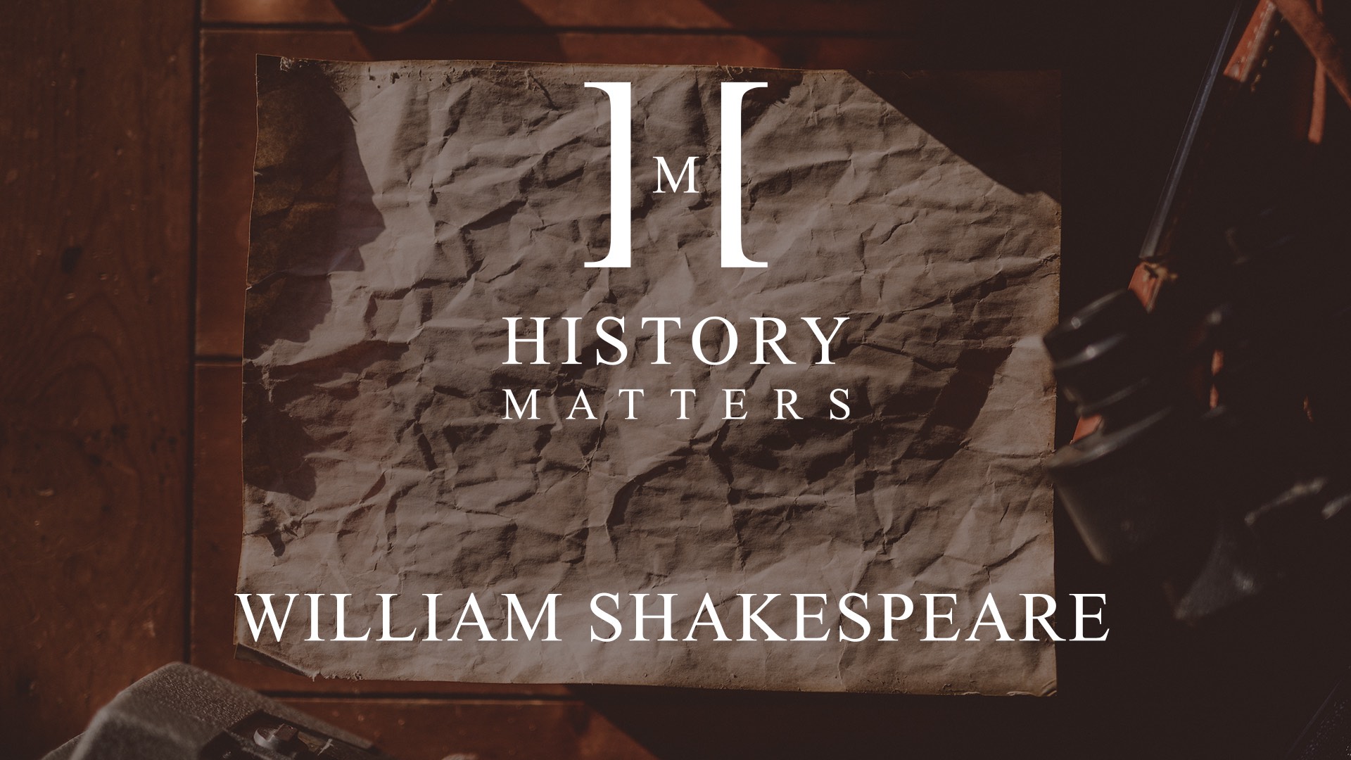 White HM William Shakespeare logo with background showing crumpled paper and binoculars on display