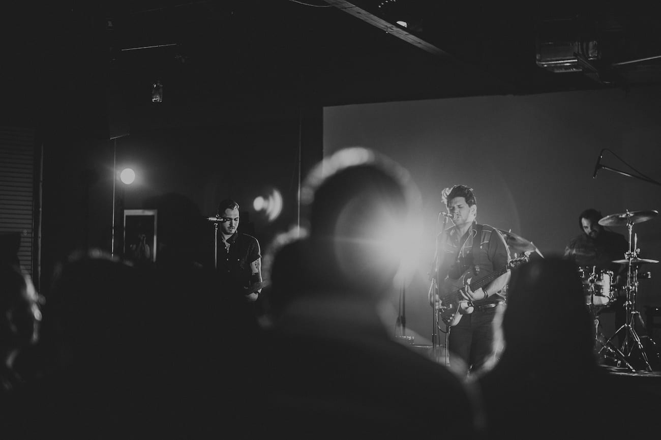 Black and white of three male musicians, a drummer and two guitarists, performing at a concert