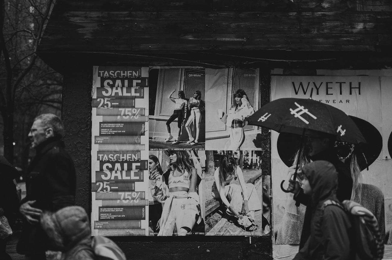 Black and white photo of a group of people walking past a bunch of billboards relating to fashion.