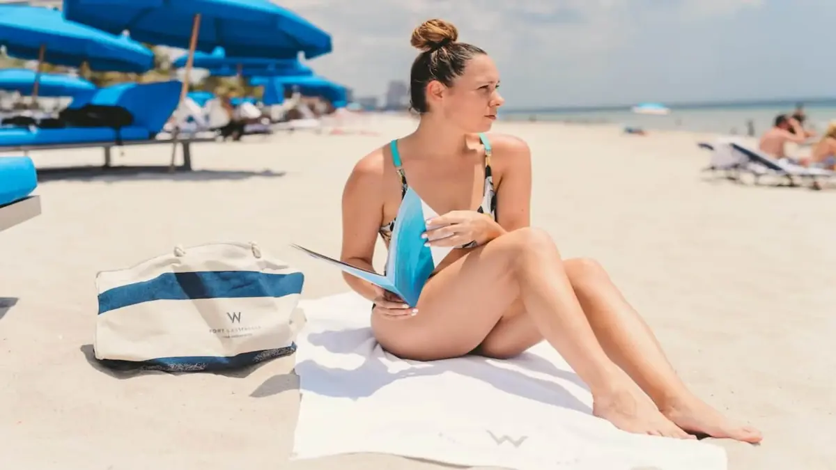 W Fort Lauderdale 12 Woman with blond hair in a bun holding a brochure sitting on a white towel on the beach looking off to the side