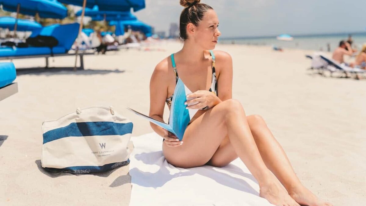 W Fort Lauderdale 12 Woman with blond hair in a bun holding a brochure sitting on a white towel on the beach looking off to the side