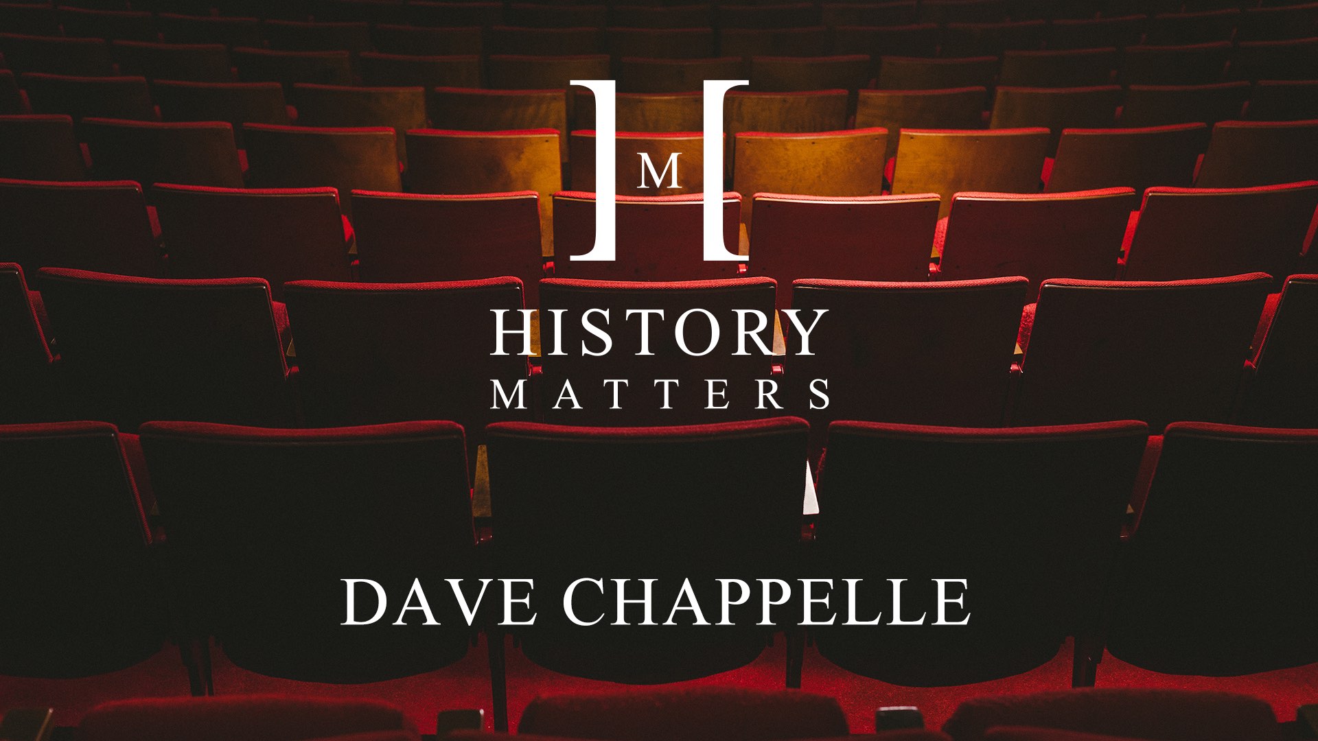 White HM Dave Chappelle logo with background showing movie seats in a movie theater