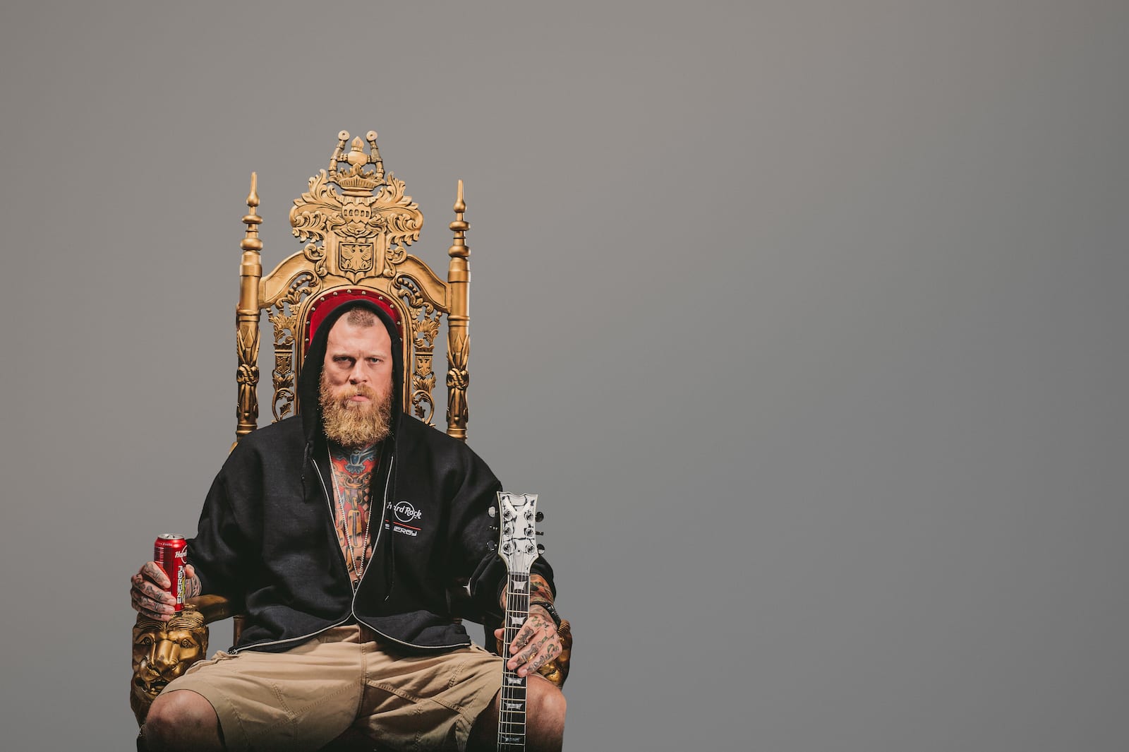 Bearded tattooed man holding Hard Rock Energy drink in one hand and a guitar in the other sitting on a throne posing for camera wearing a black hooded jacket