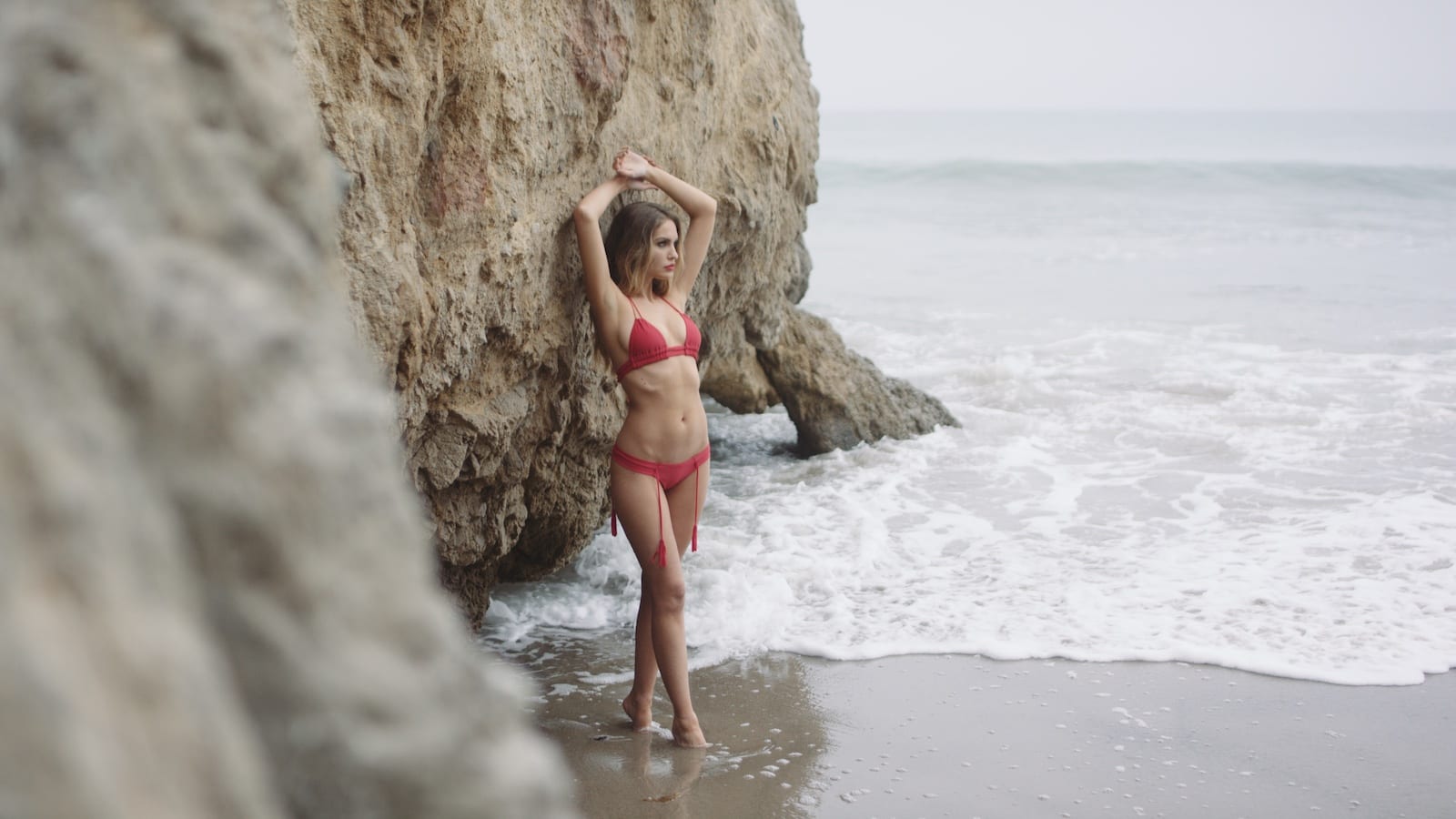 Montce Swim Ad 2.28.1 Woman with long blond hair wearing a red bikini posing for camera on a beach standing in the water with her arms over her head leaning against a rocky outcrop