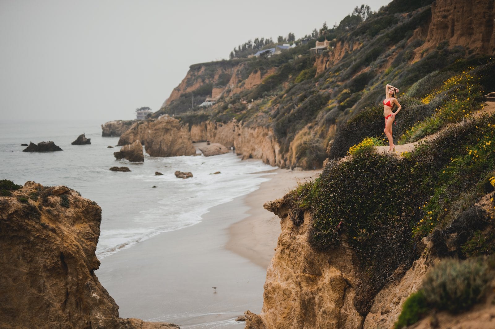 International Marketing for Montce Swim ReachKini 6 Woman with long brown hair wearing a red bikini posing for the camera from on top of a rocky outcrop by a beach