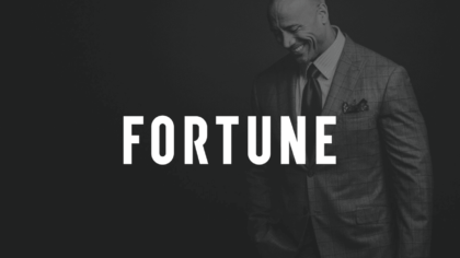 Black And White Of White Fortune Logo With Dwayne "The Rock" Johnson With Fortune Magazine In The Background Posing For Camera Looking Down And Smiling