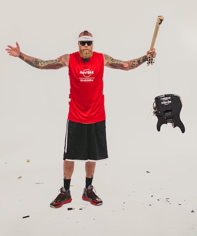 Bearded tattooed man wearing a red Hard Rock Energy jersey and black shorts holding part of a guitar with other guitar parts flying around