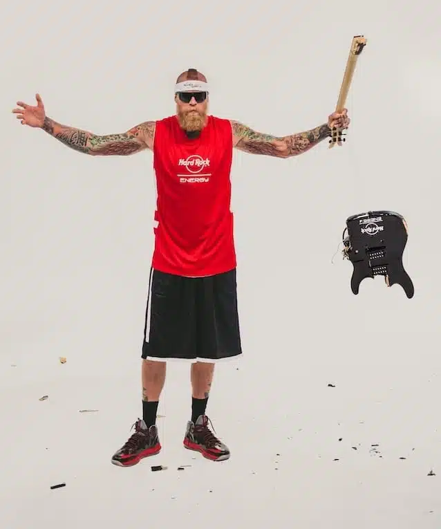 Hard Rock Energy 18 Bearded tattooed man wearing a red Hard Rock Energy jersey and black shorts holding part of a guitar with other guitar parts flying around