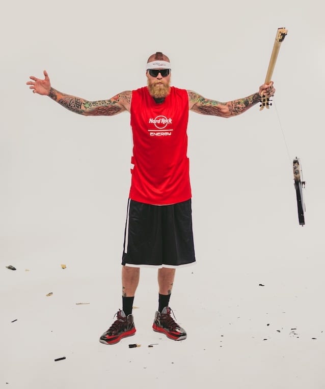 Bearded tattooed man wearing a red Hard Rock Energy jersey and black shorts holding part of a guitar with other guitar parts flying around