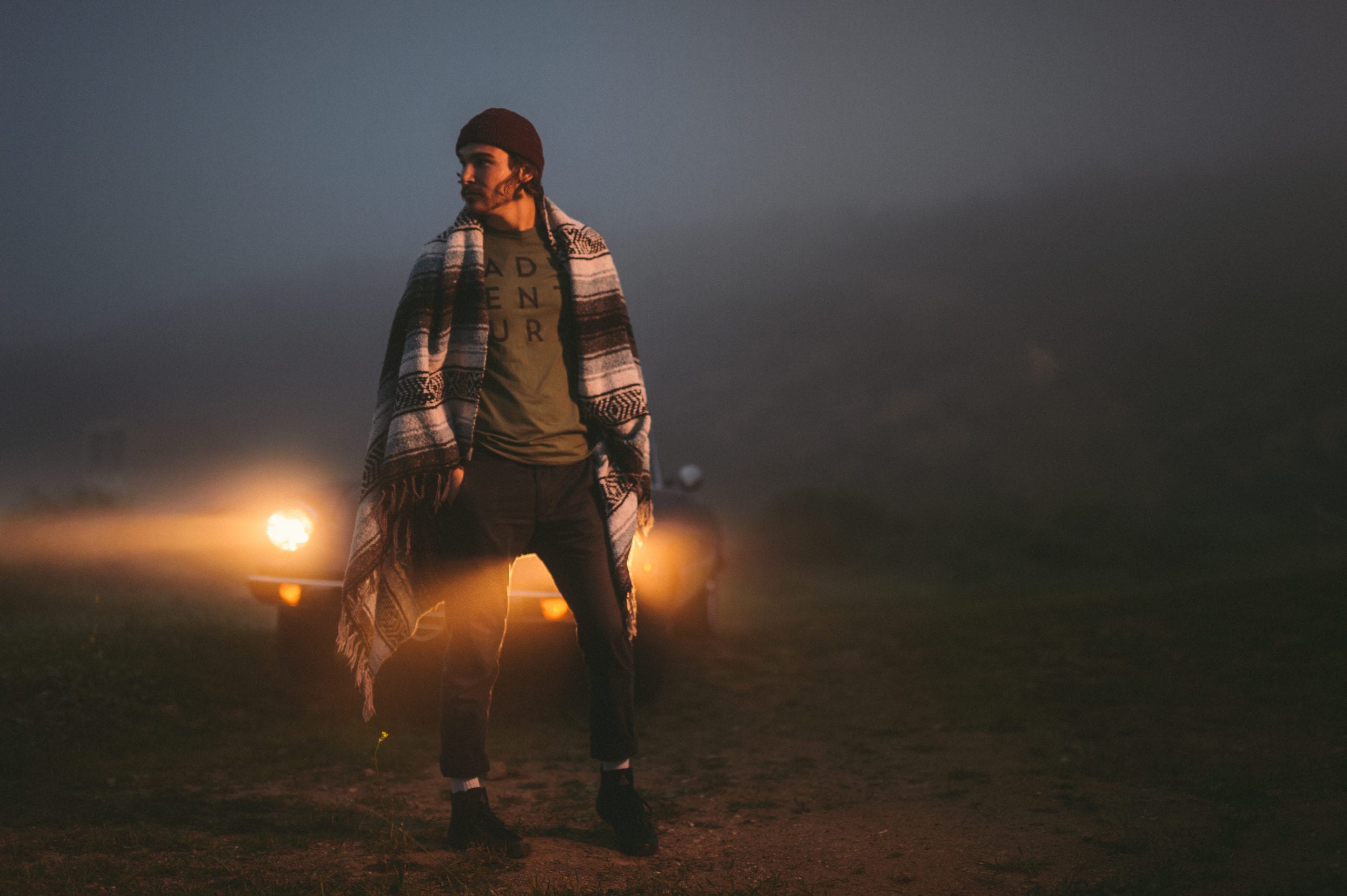 Professional Photography Services for retail and clothing brands The Shop Man wearing knitted cap and Mexican blanket posing for the camera looking off camera in front of a car with headlights on at dusk