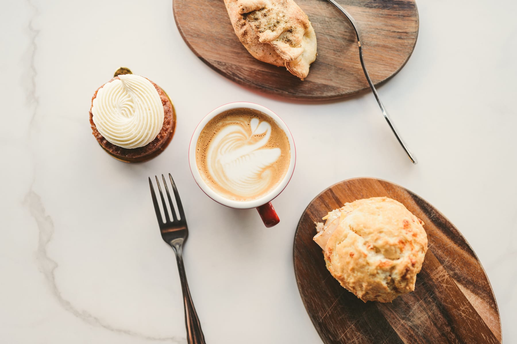 Brew Urban Cafe Spread of pastries on wooden plates, muffin with white frosting and red cup of cappuccino on display on marble table with two forks