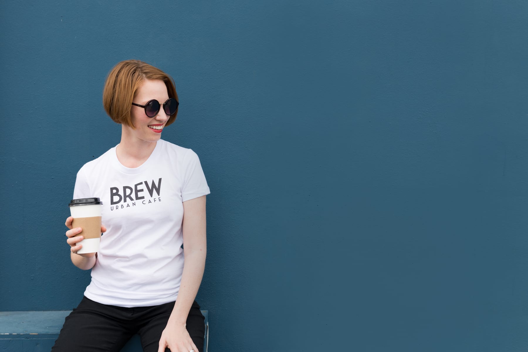 Brew Urban Cafe Woman with short hazel hair wearing sunglasses, white Brew Urban Cafe tshirt holding a drink smiling and posing for camera
