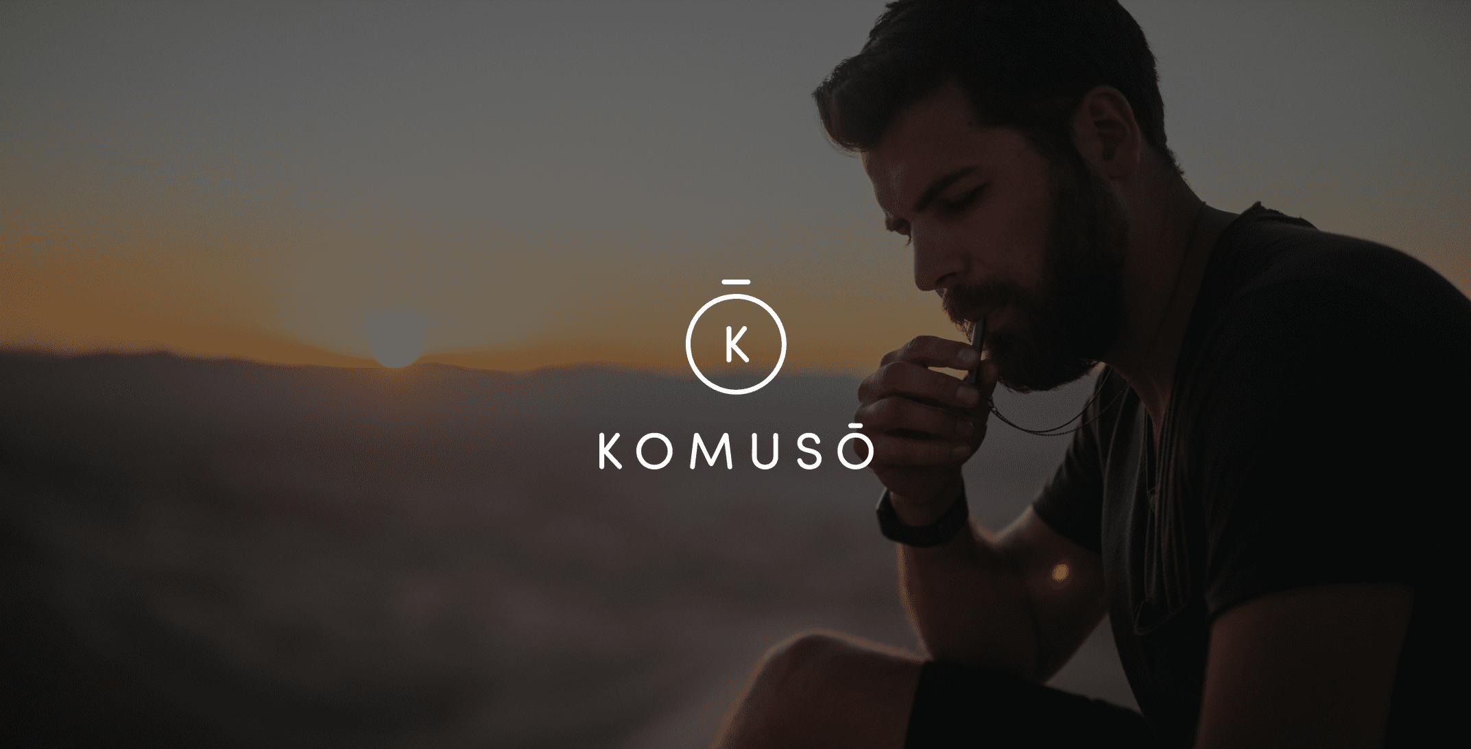 White Komuso logo with side profile backdrop of man with a beard and short hair blowing a whistle with a valley and the sun in the background