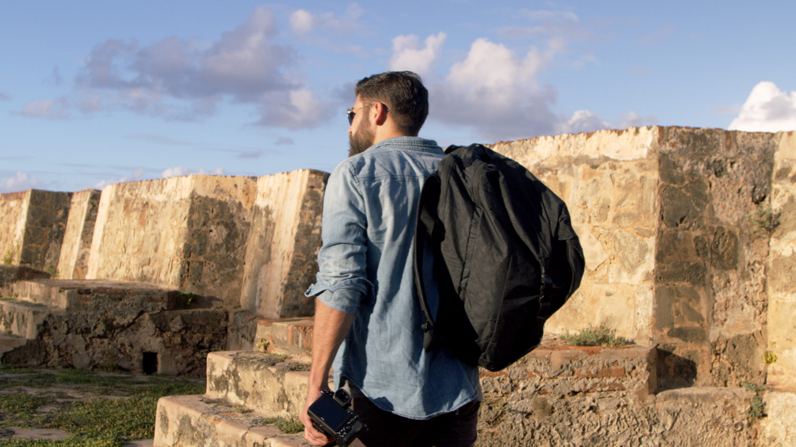 Blackmile Closeup view from behind of a man wearing a jean shirt and black backpack holding a camera looking out of the stone walls