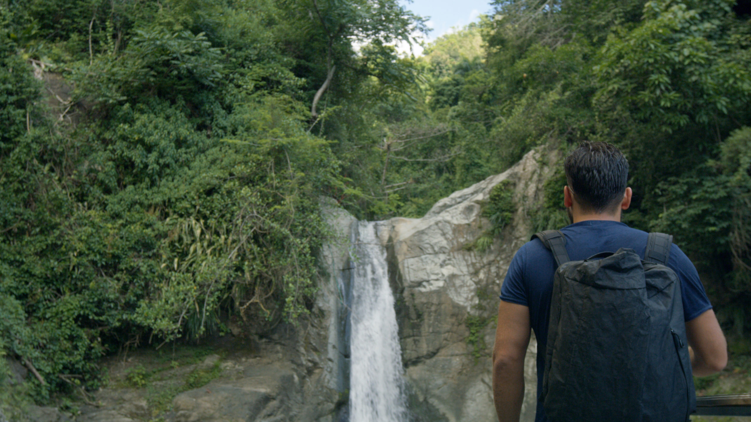 Blackmile Man wearing a backpack wearing a dark navy tshirt looking at a waterfall