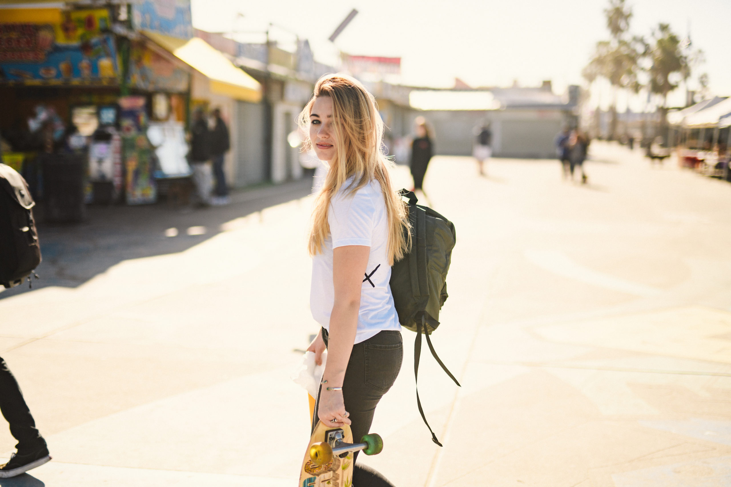 Artistic Bloggers Venice Beach Uncreative Blog Side profile of a woman with long blond hair posing for the camera wearing a green backpack and carrying a skateboard