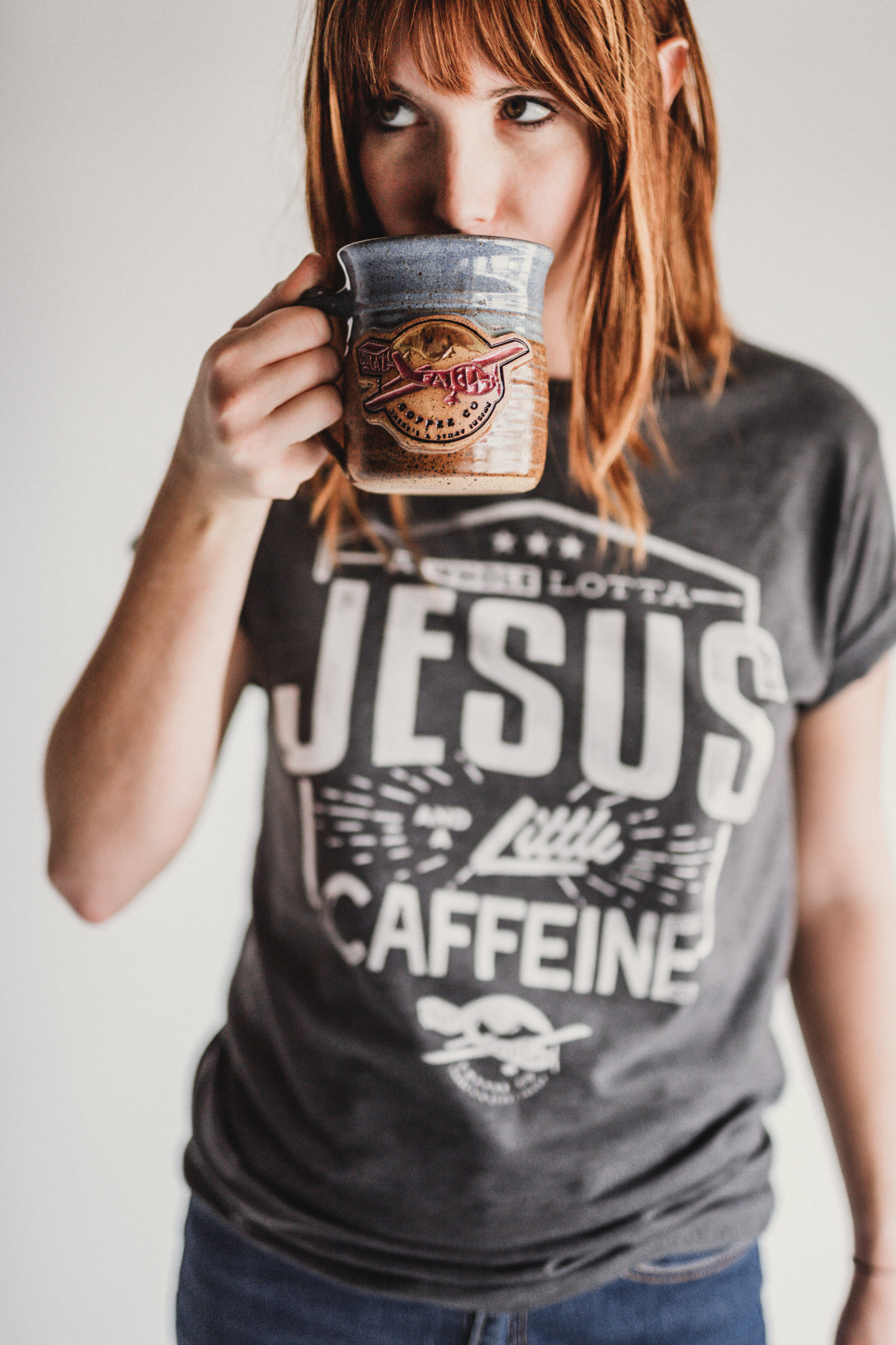 JSM Woman wearing a gray t shirt that says A Whole Lotta Jesus and a Little Caffeine from Crazy Faith drinking some coffee from a gray coffee cup