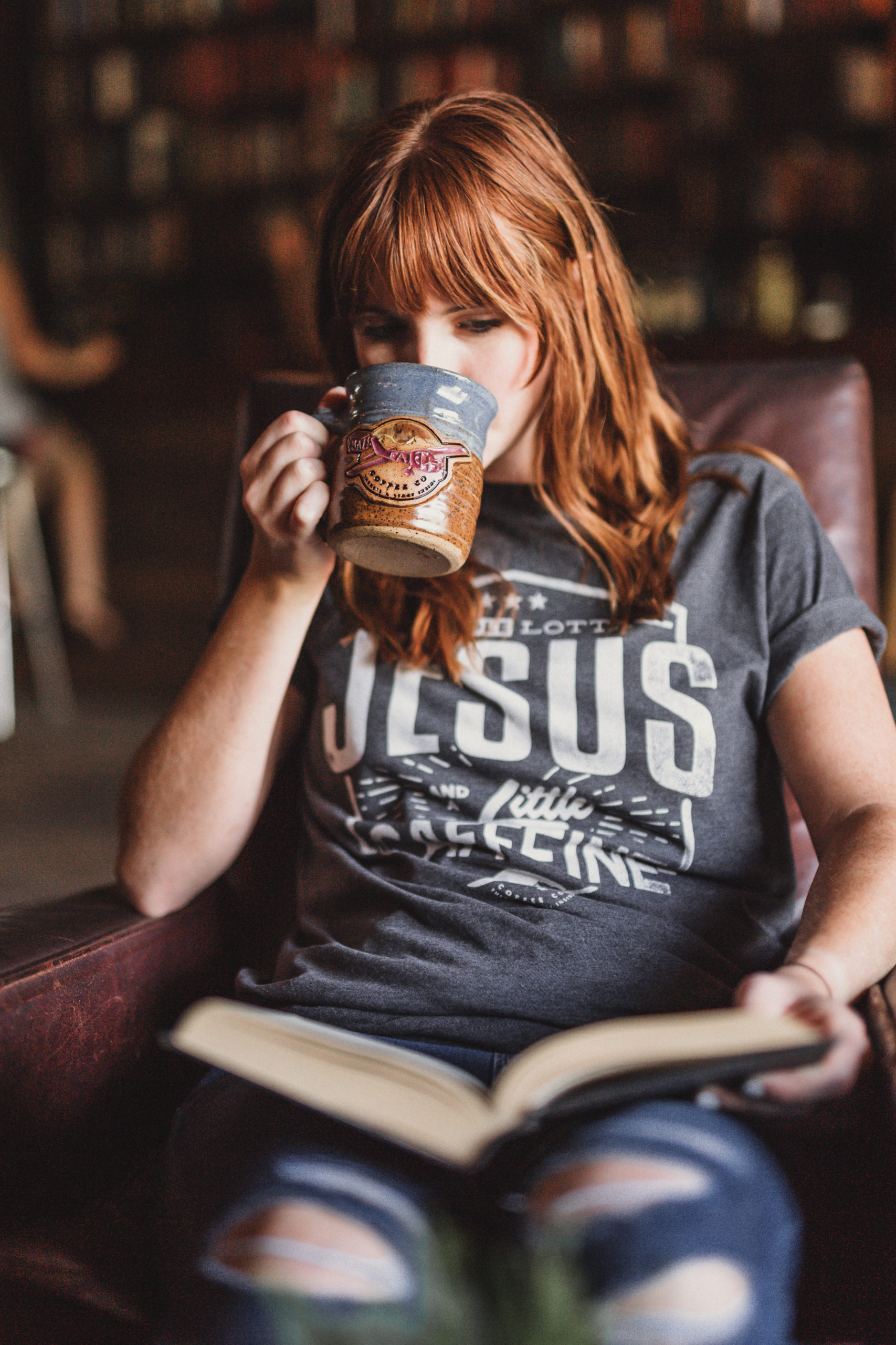 JSM Woman wearing a gray t shirt that says A Whole Lotta Jesus and a Little Caffeine from Crazy Faith drinking some coffee from a brown and gray coffee cup and reading a book