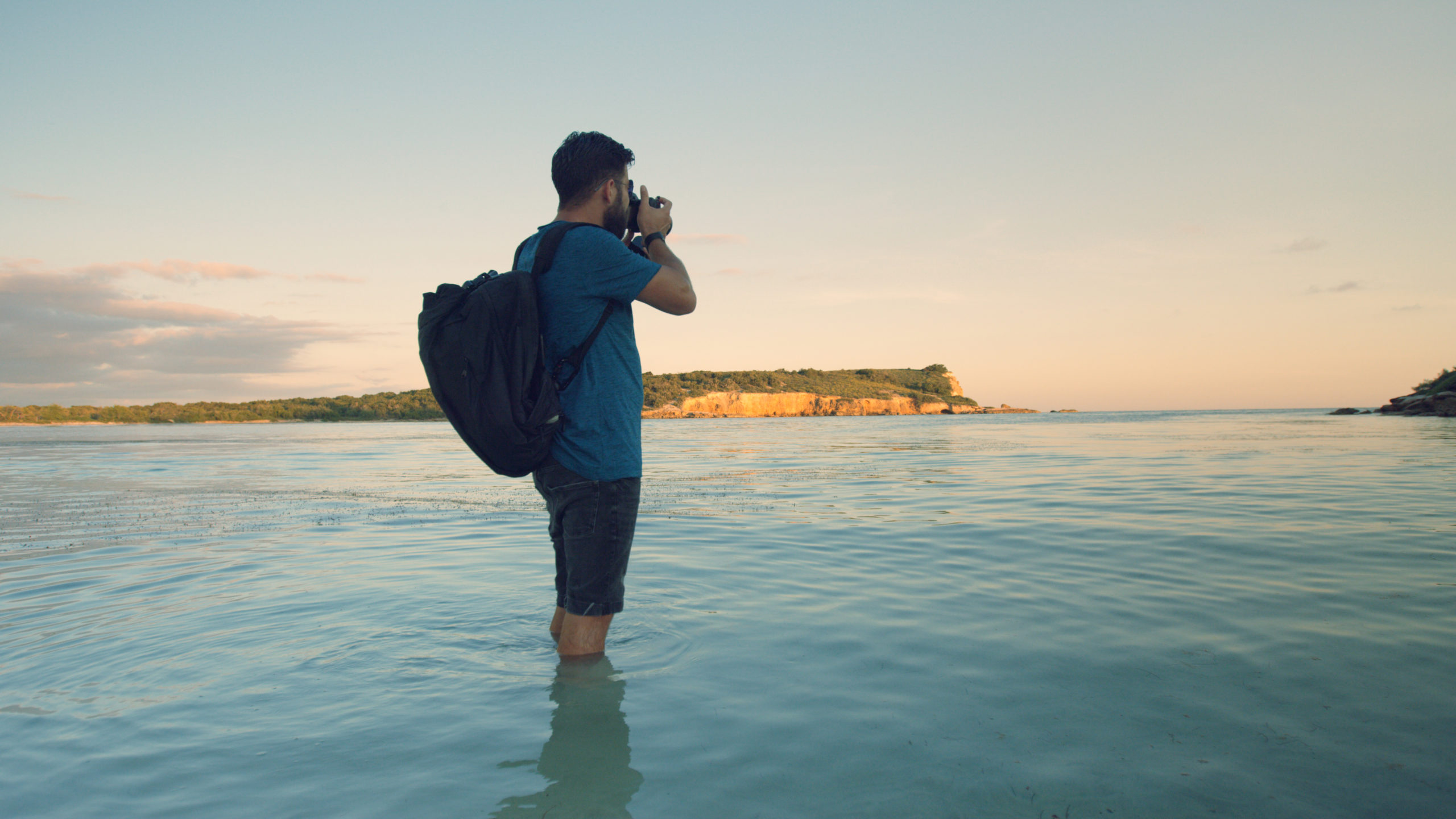 Man talking a picture of an island standing in the water wearing a backpack