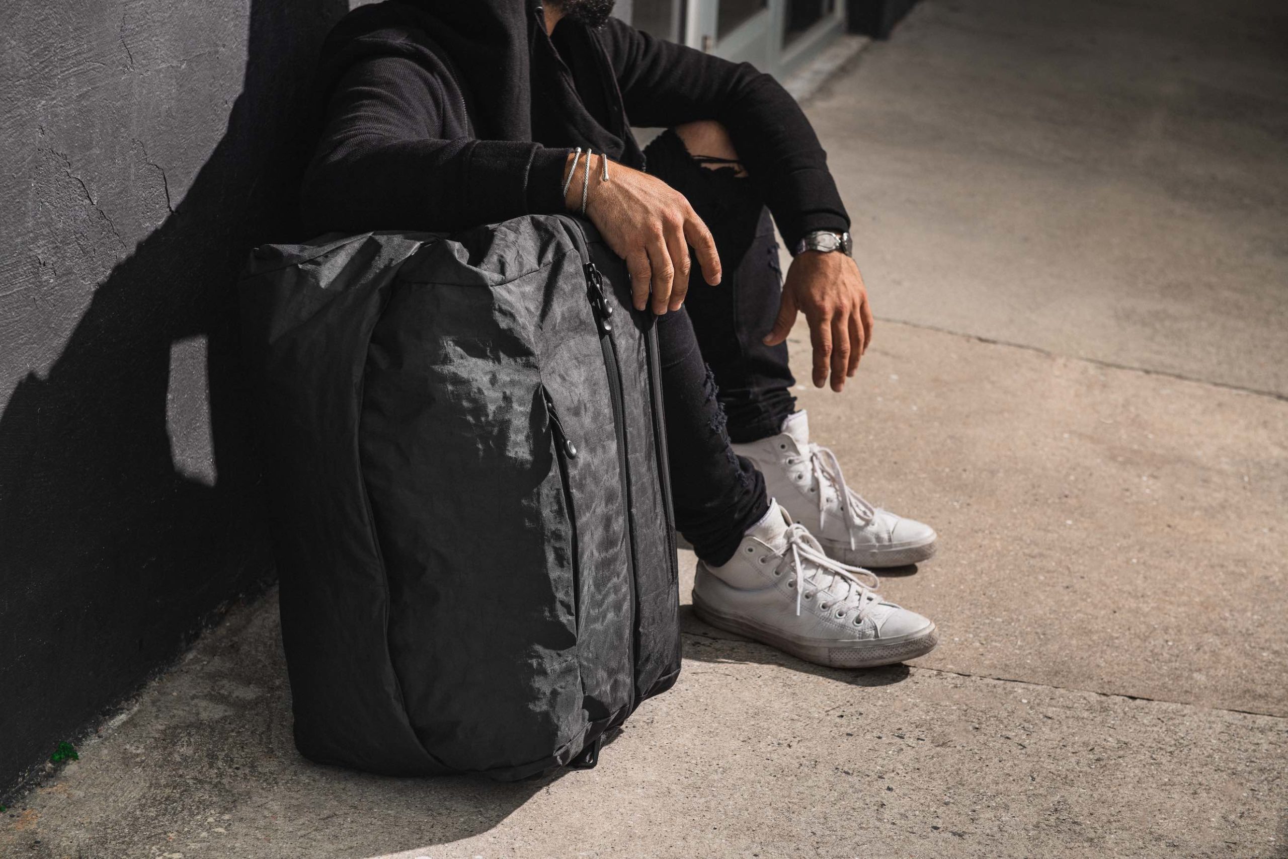 Side profile of a man wearing white shoes and black jeans with a black bag next to him sitting on the floor