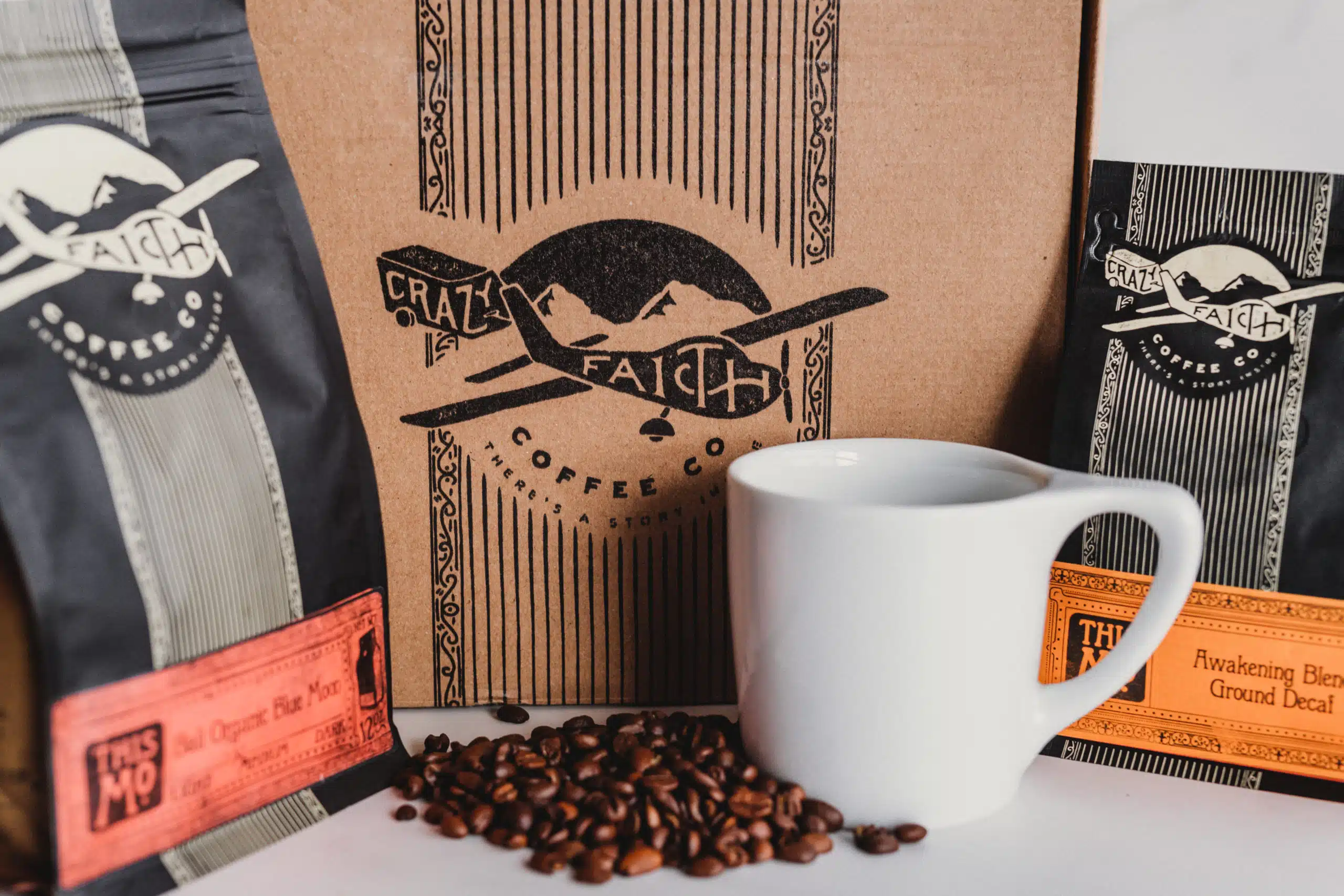 Crazy Faith Coffee 24 Packages of Bali Organic Blue Moon and Awakening Blend Ground Decaf coffee beans with a box in the background as well as coffee beans and white coffee cup