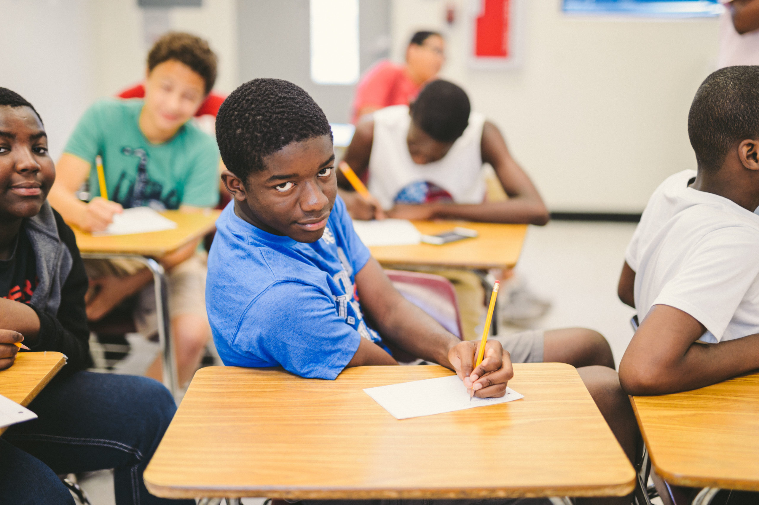 Group of young adults in a classroom with one African American student wearing a blue shirt posing for the camera holding a pencil on a piece of paper