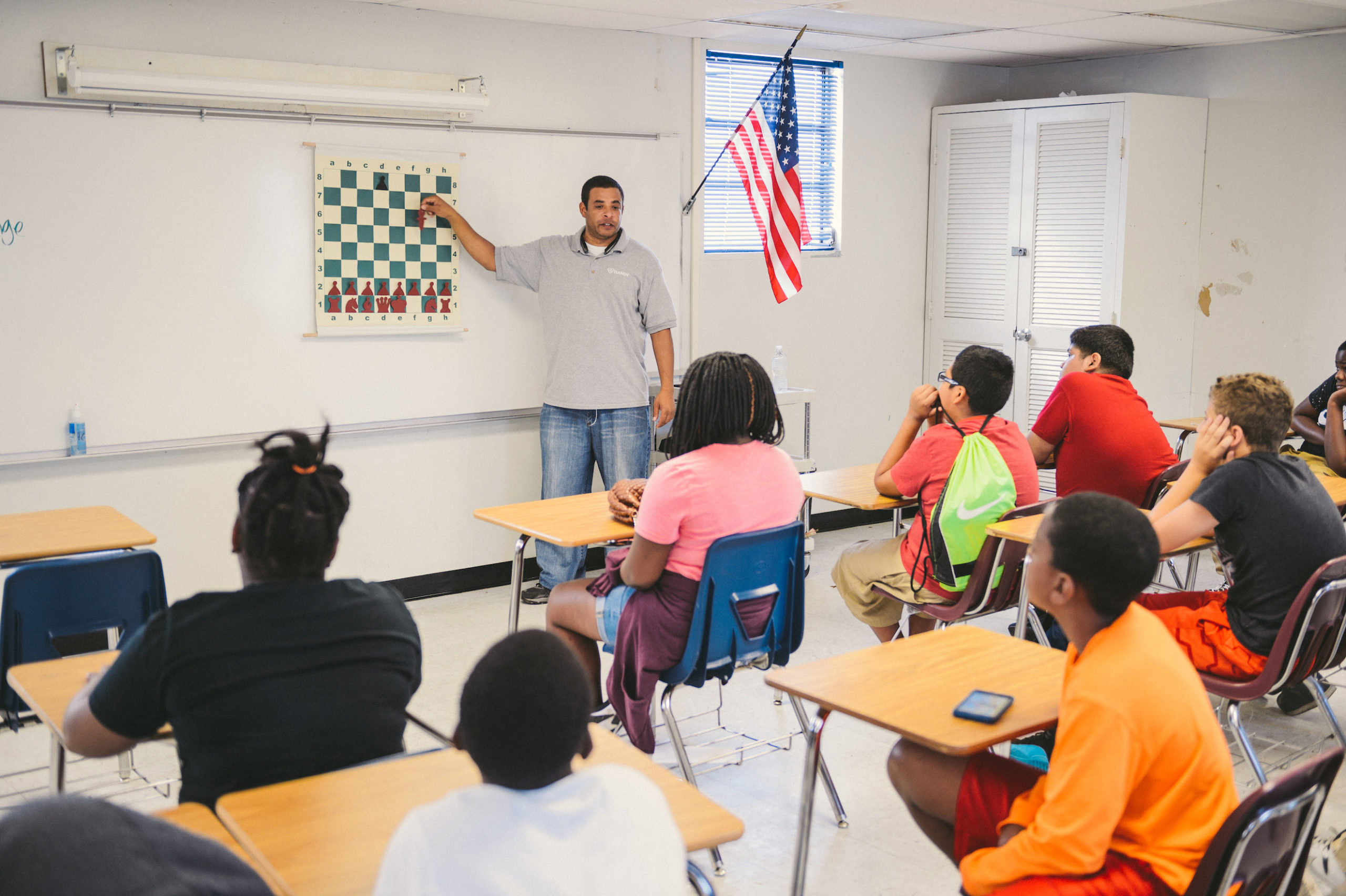 Teacher showing how to play a game of chess to a class of young adults sitting at their desks
