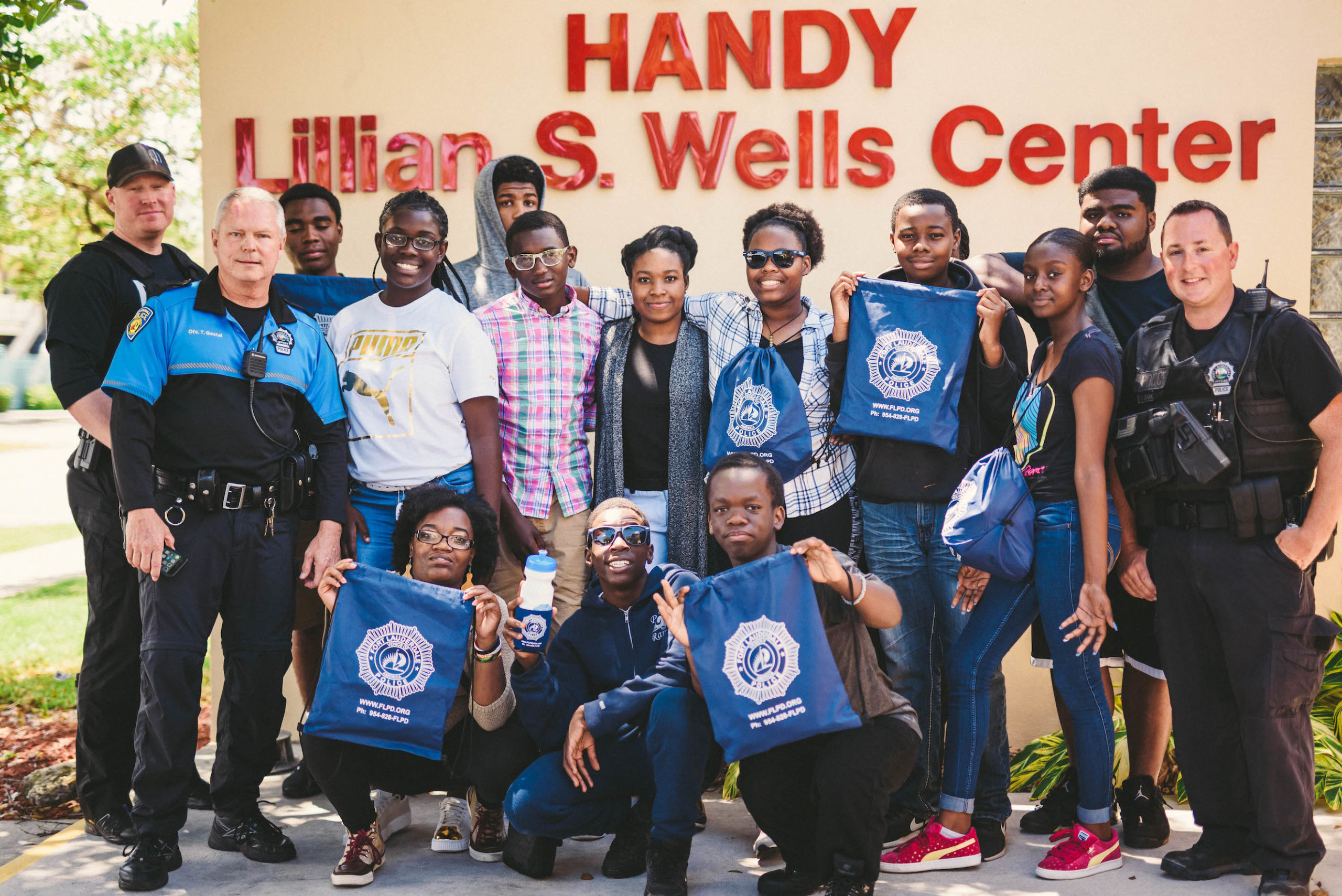 Young adults with two police officers posing in front of Handy Lillian S. Wells Center sign with some holing police logo bags and water bottle