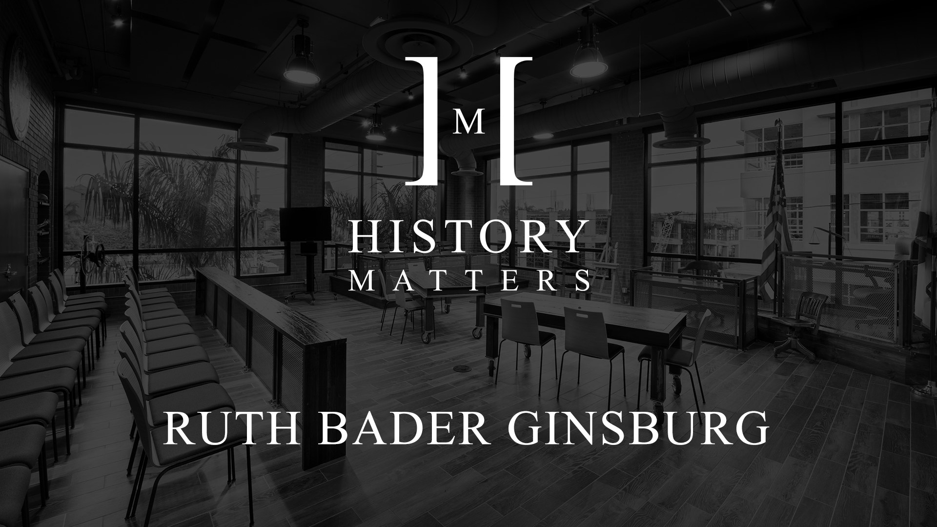 White HM Ruth Bader Ginsburg logo with dimmed black and white background of a classroom with chairs, tables and an American Flag logo with background