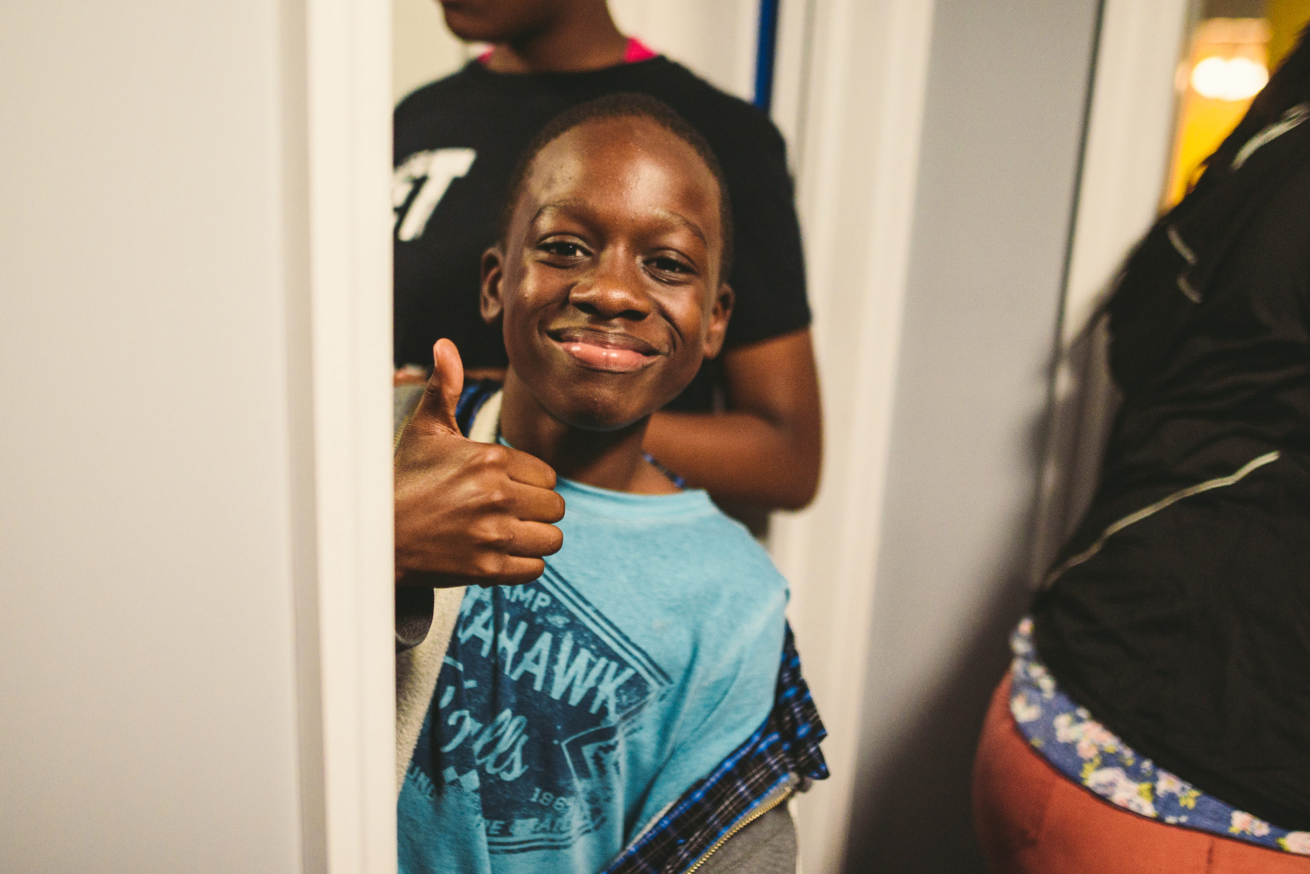 Handy Nonprofit African American boy wearing a light blue shirt with logo giving a thumbs up and smiling surrounded by people
