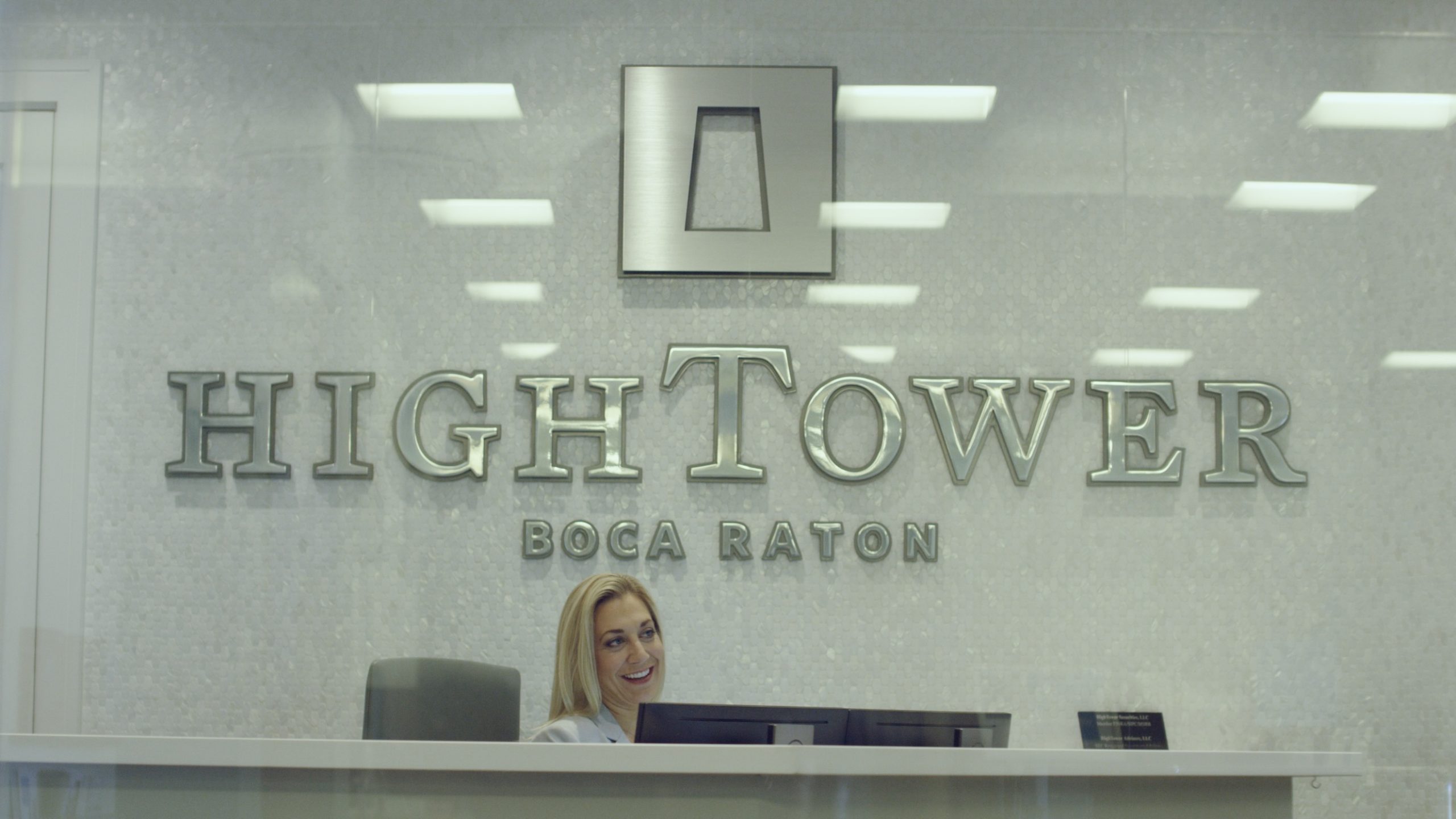 Hightower Financial Advisors Hightower Boca Raton logo in a lobby of the company with a secretary sitting at her desk smiling for the camera looking off to the side