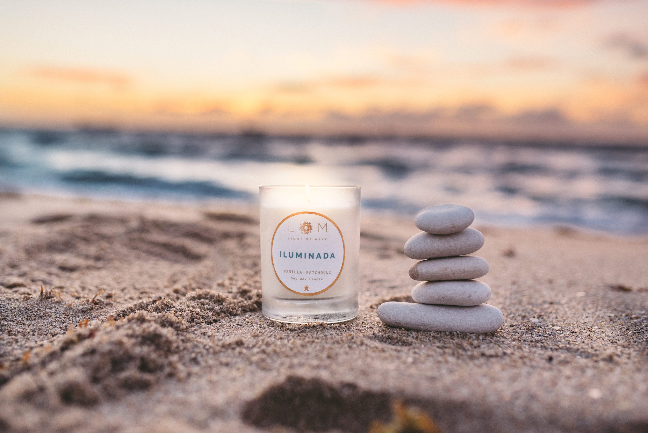 Closeup of Light of Mine soy wax candle on a beach with a stack of stones nearby.