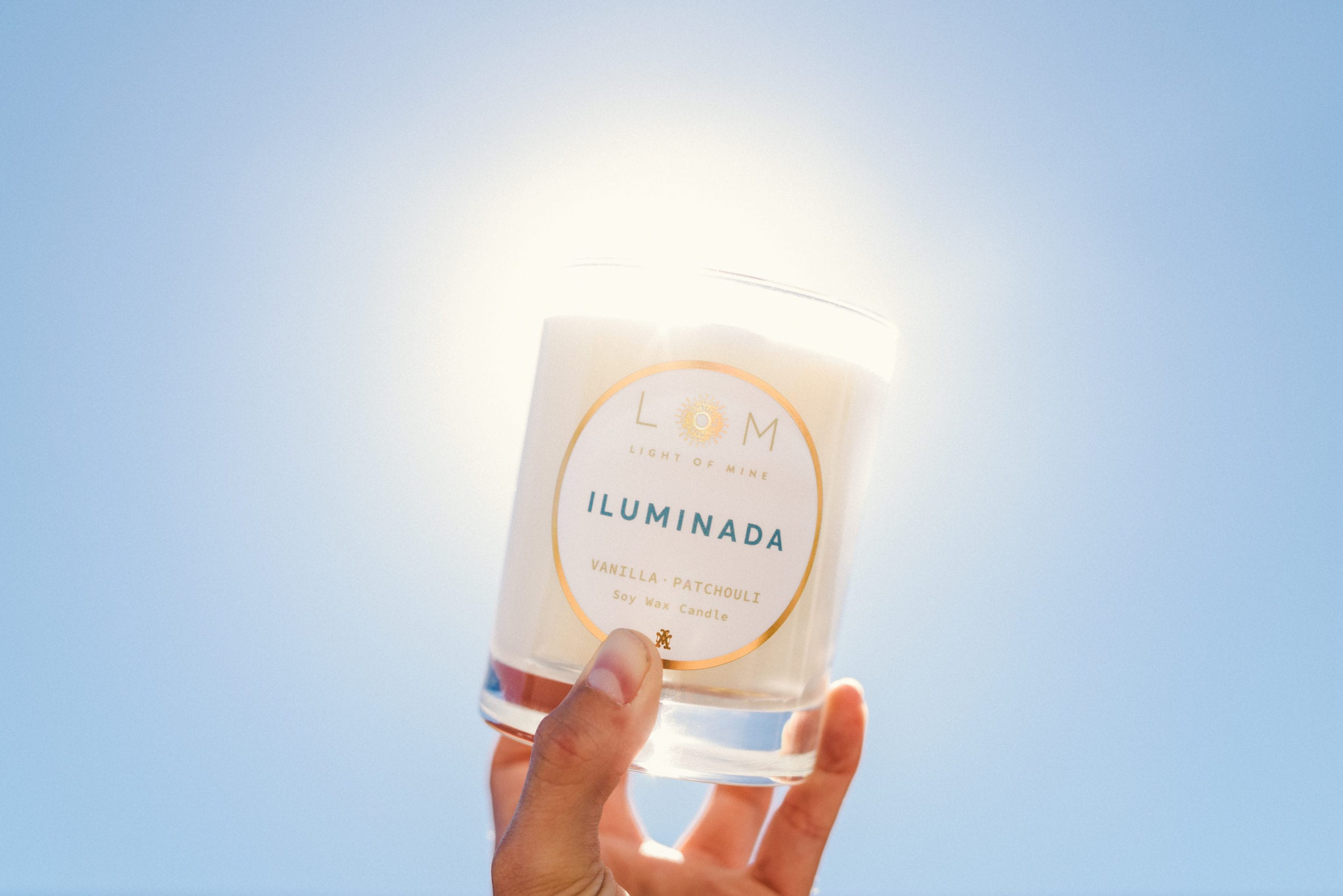 Closeup of hand holding up a LOM Light of Mine Iluminada Vanilla Patchouli candle against the sun
