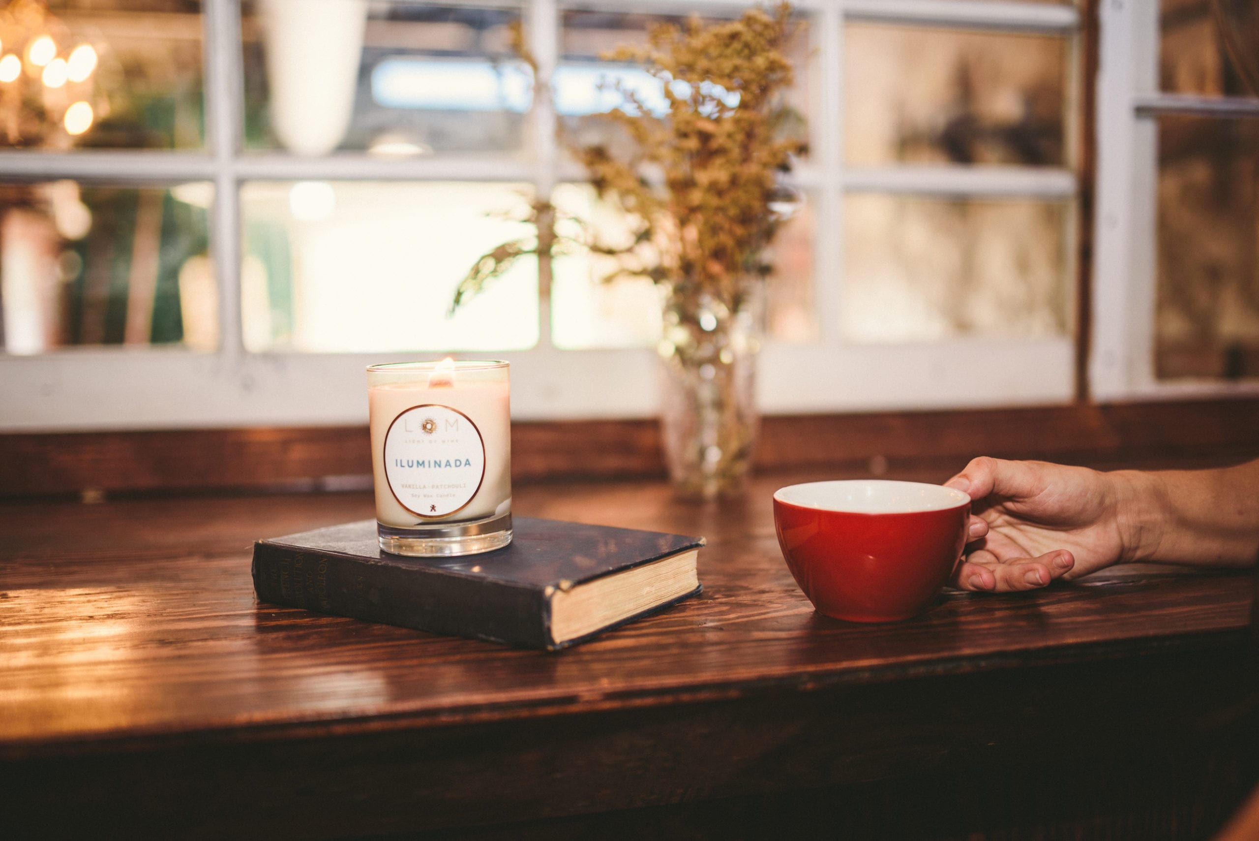 Closeup of a person holding a red coffee mug next to a book with a LOM Light of Mine Iluminada candle on a wooden table