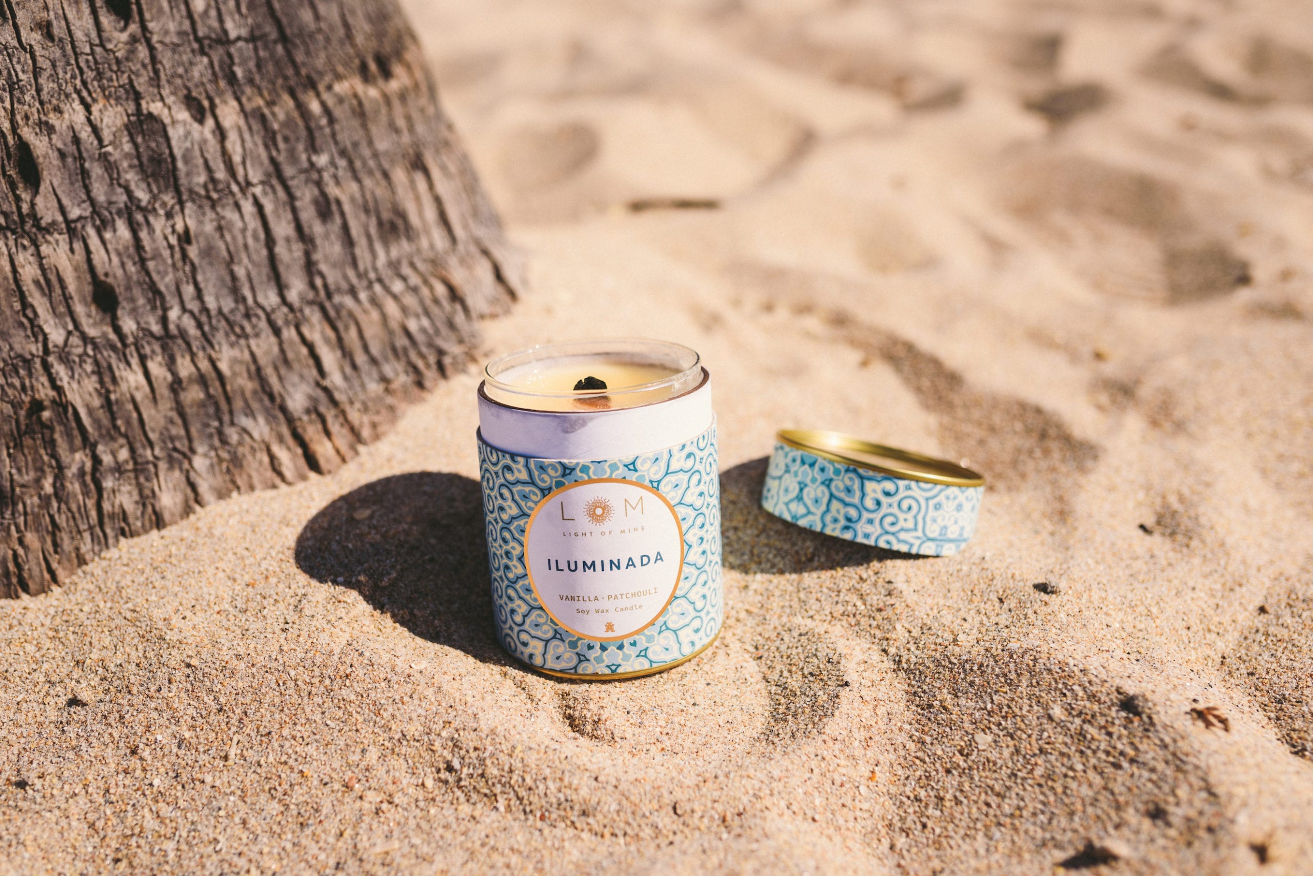LOM Light of Mine Vanilla Patchouli candle in the sand next to lid with palm tree trunk in background