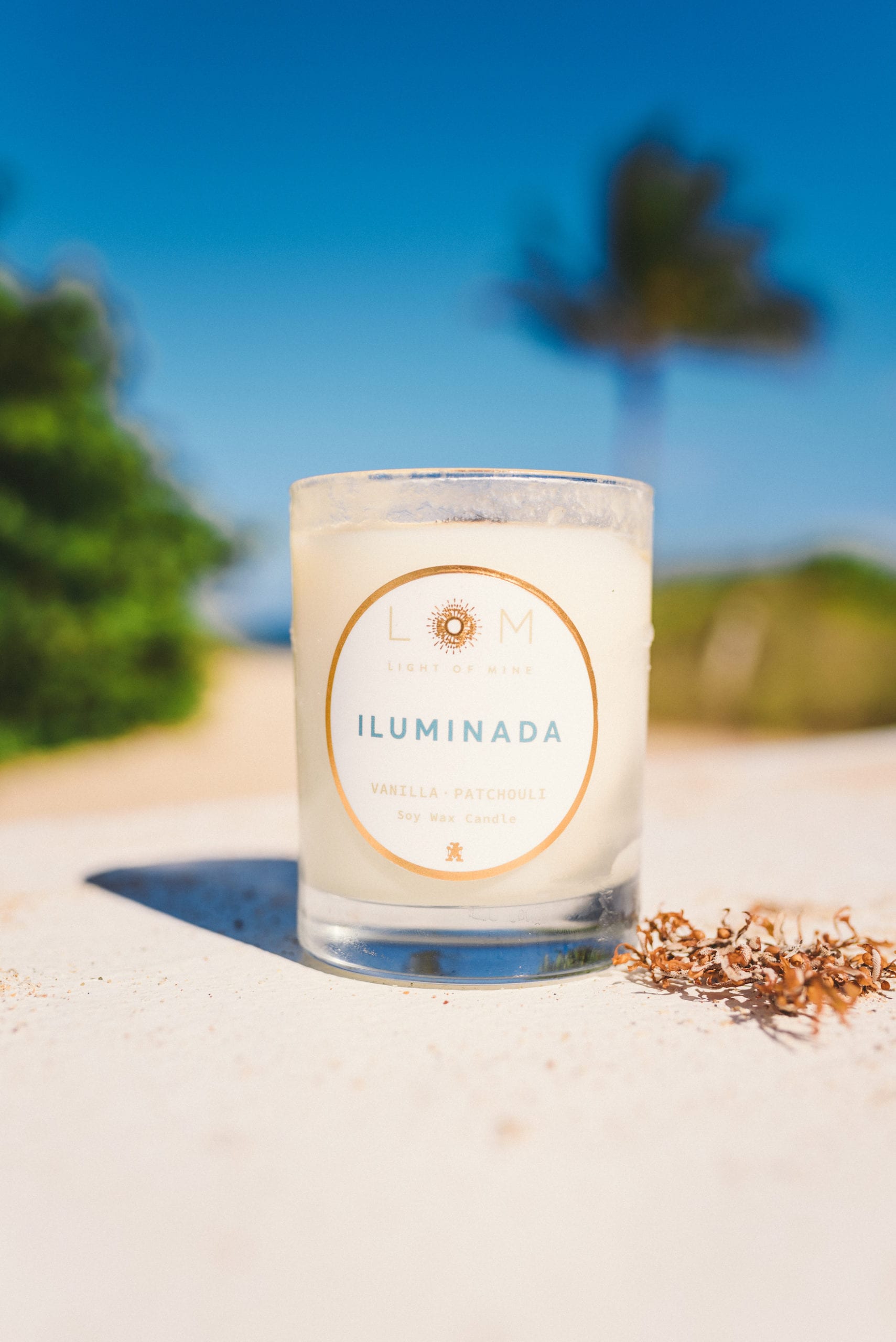 Closeup of LOM Light of Mine Iluminada Vanilla Patchouli candle on a table against a beach and palm tree backdrop