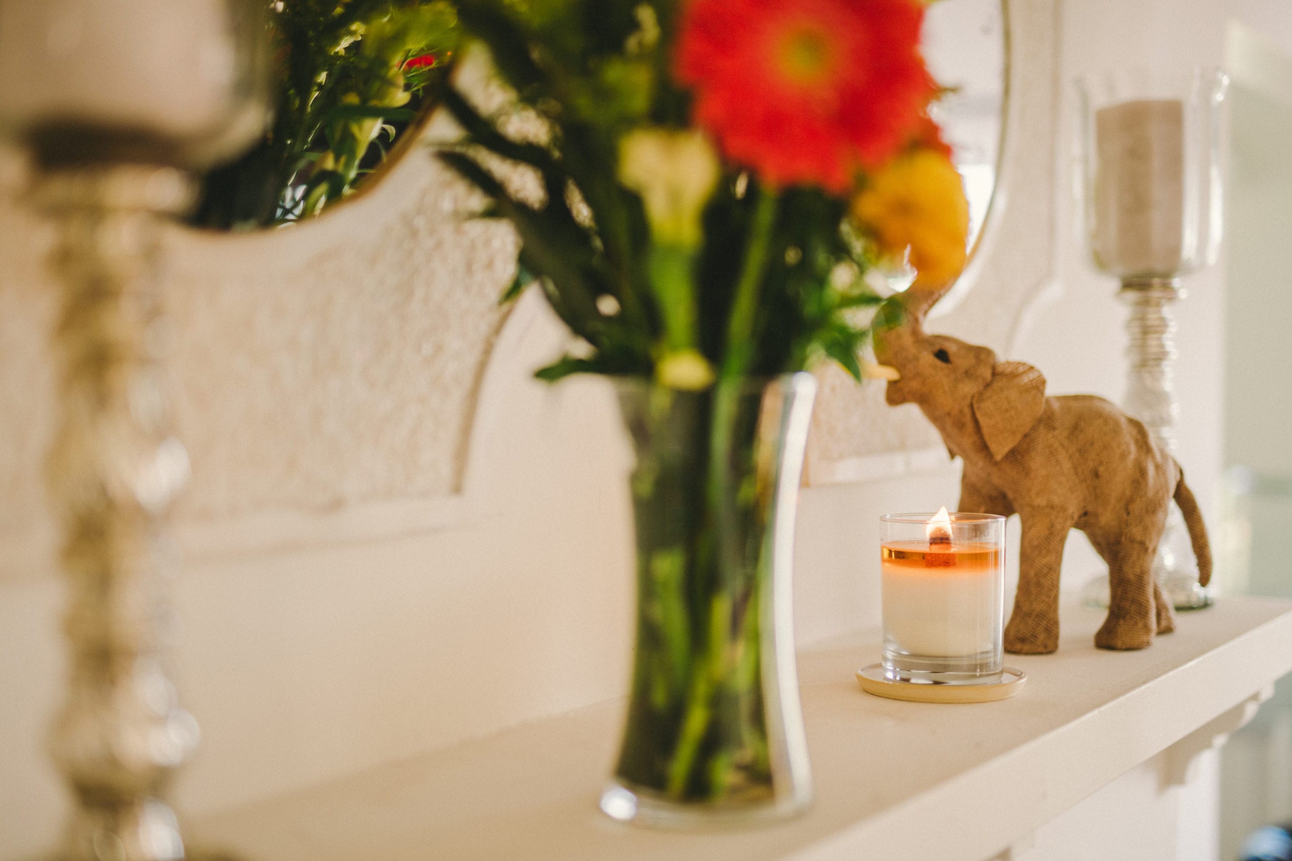 JSM Candle with orange liquid on shelf in between elephant art in the background and vase of flowers in the foreground