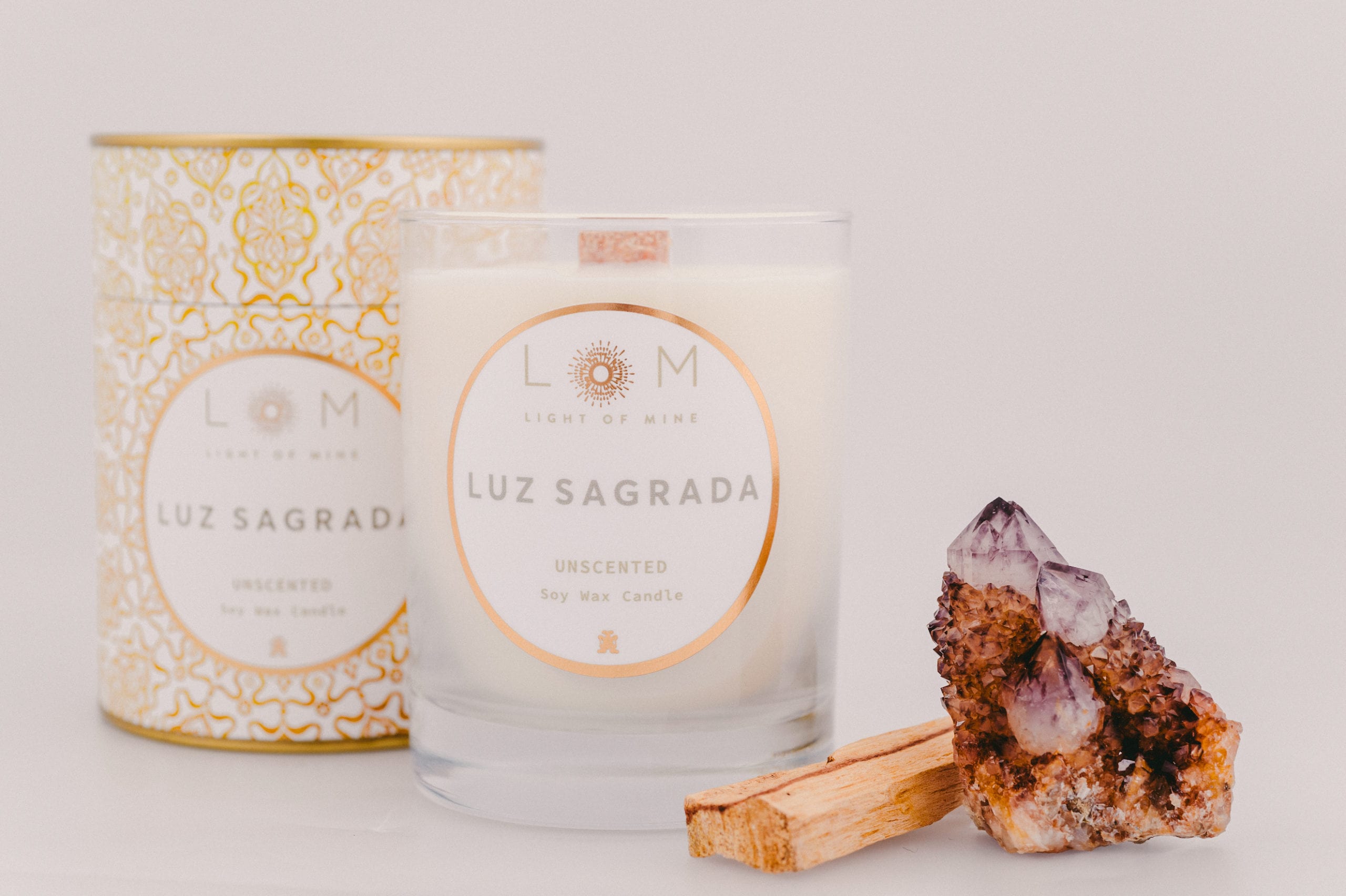 Two Light of Mine Luz Sagrada brand unscented soy wax candles next to a piece of wood and large quartz stone