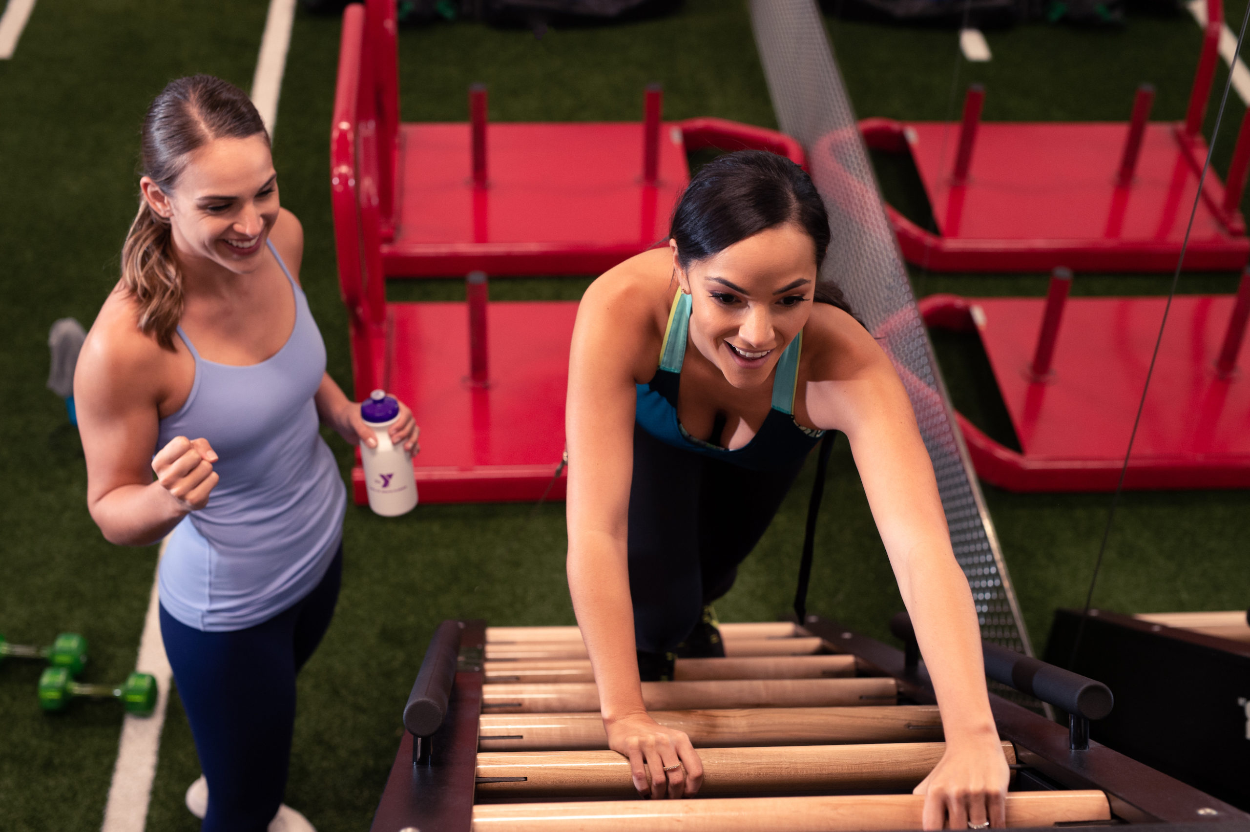 YMCA Overhead view of a woman climbing an exercise ladder with another woman cheering her on with both smiling