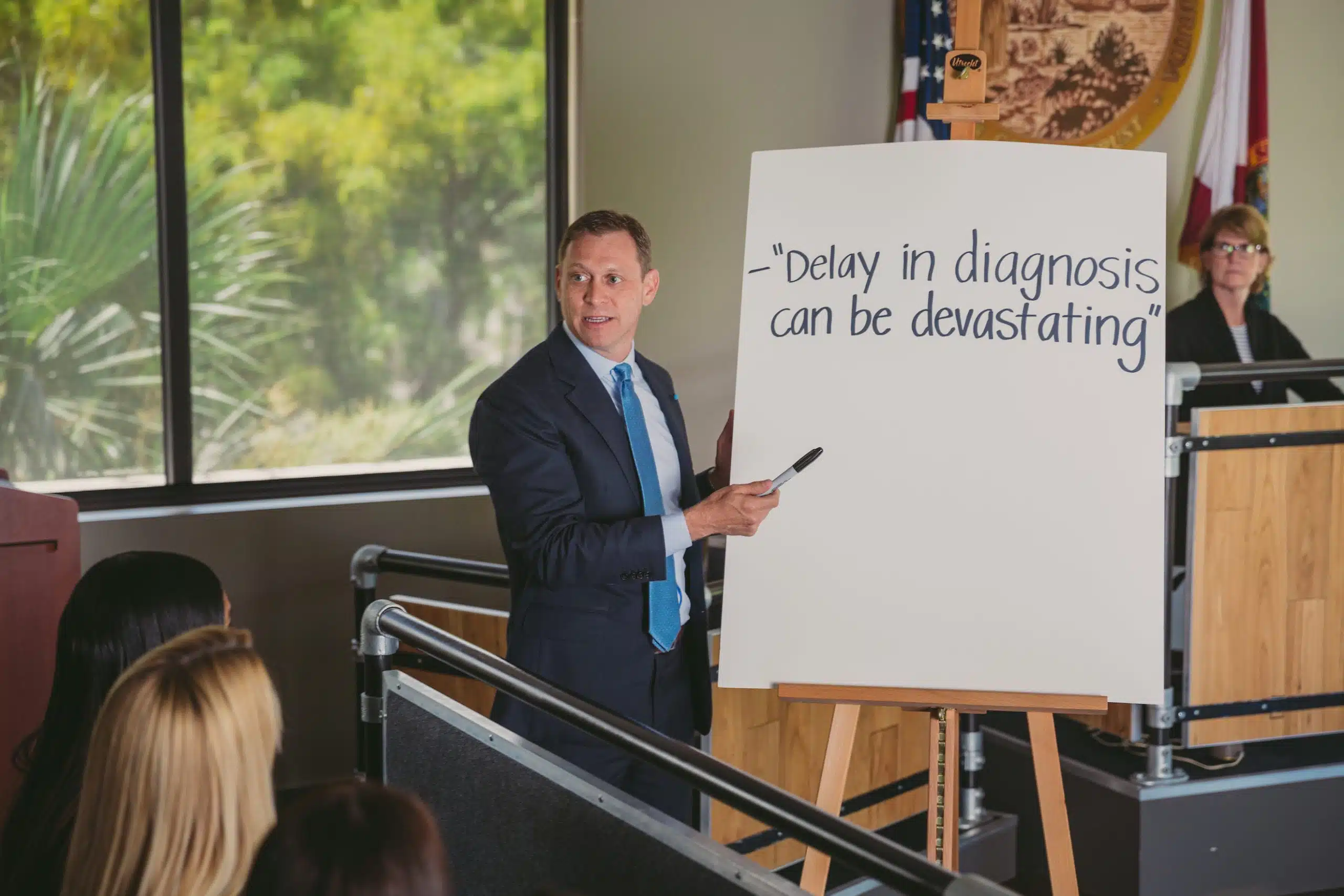 Male attorney in a blue shirt and blue tie talking to a jury next to a white board on a stand that says Delay in diagnosis can be devasting with the judge in the background in a courtroom