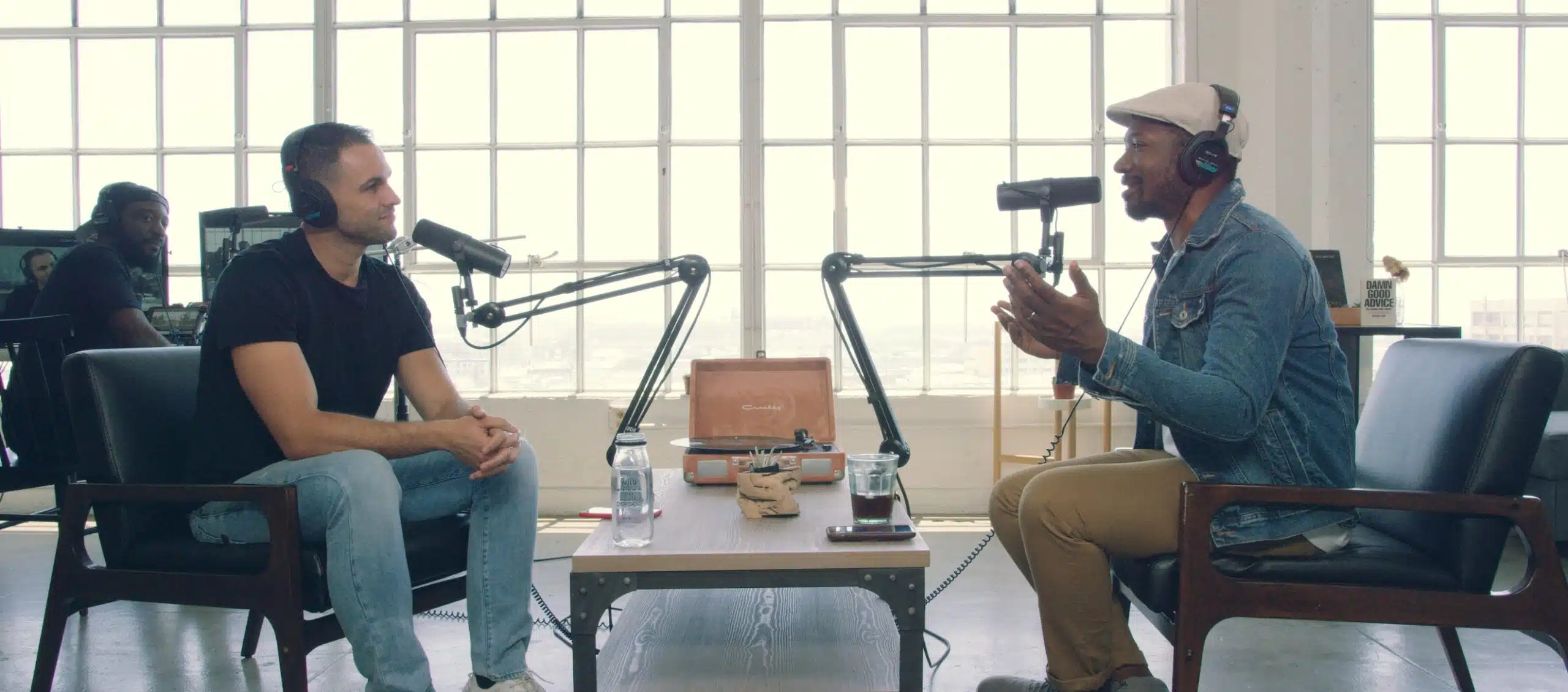 Side profile of an Joshua with a beard wearing headphones and a blue jean jacket smiling and talking in a microphone to Pavvlo Zengo along with another man listening to him wearing headphones