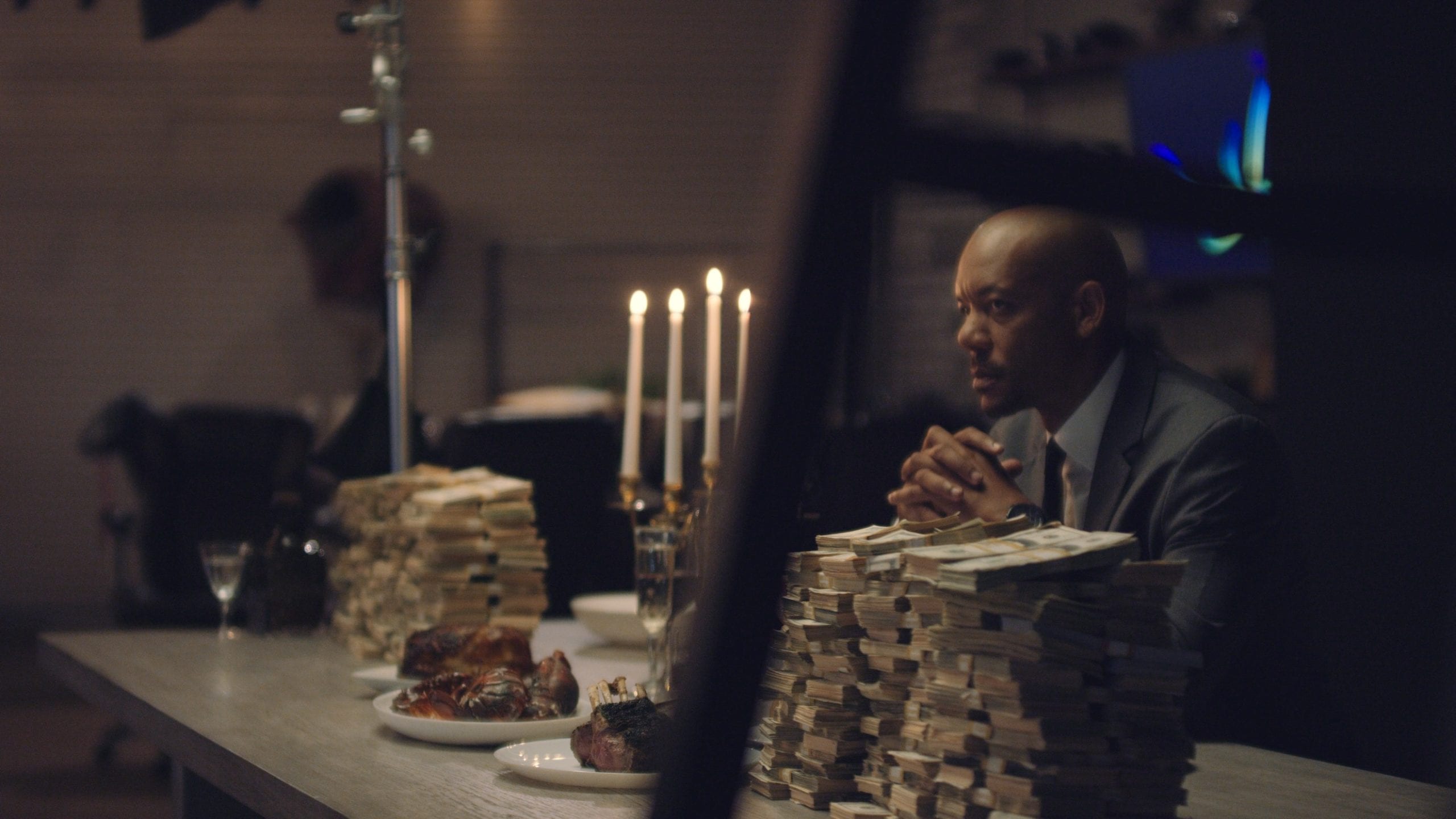 Becoming Immortal A New Original TV Series Side profile of African American man sitting at a table with food, drinks, a candelabra and stacks of cash