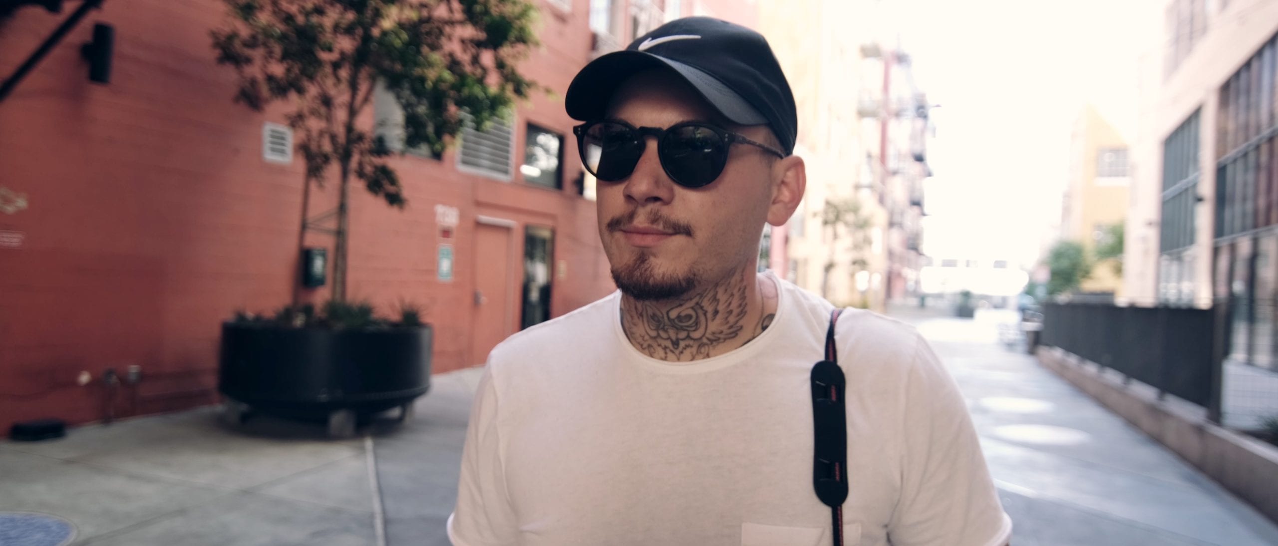 We're live with Dan Perri on Uncreative Radio EP 116 Dan with tattoos on his neck wearing a black Nike cap with white logo and sunglasses walking down an alley