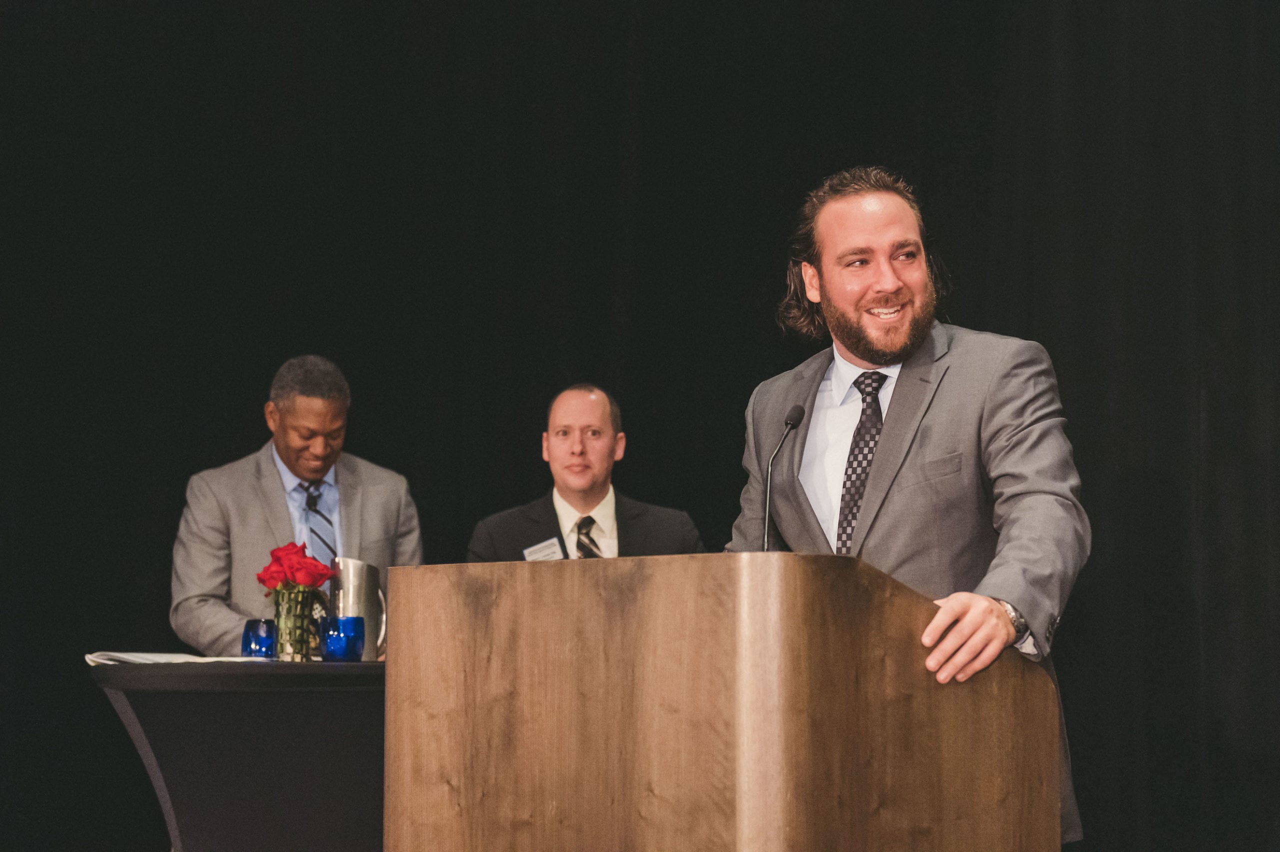 Man in a gray suit with a brown tie talking to an audience with two men in the background